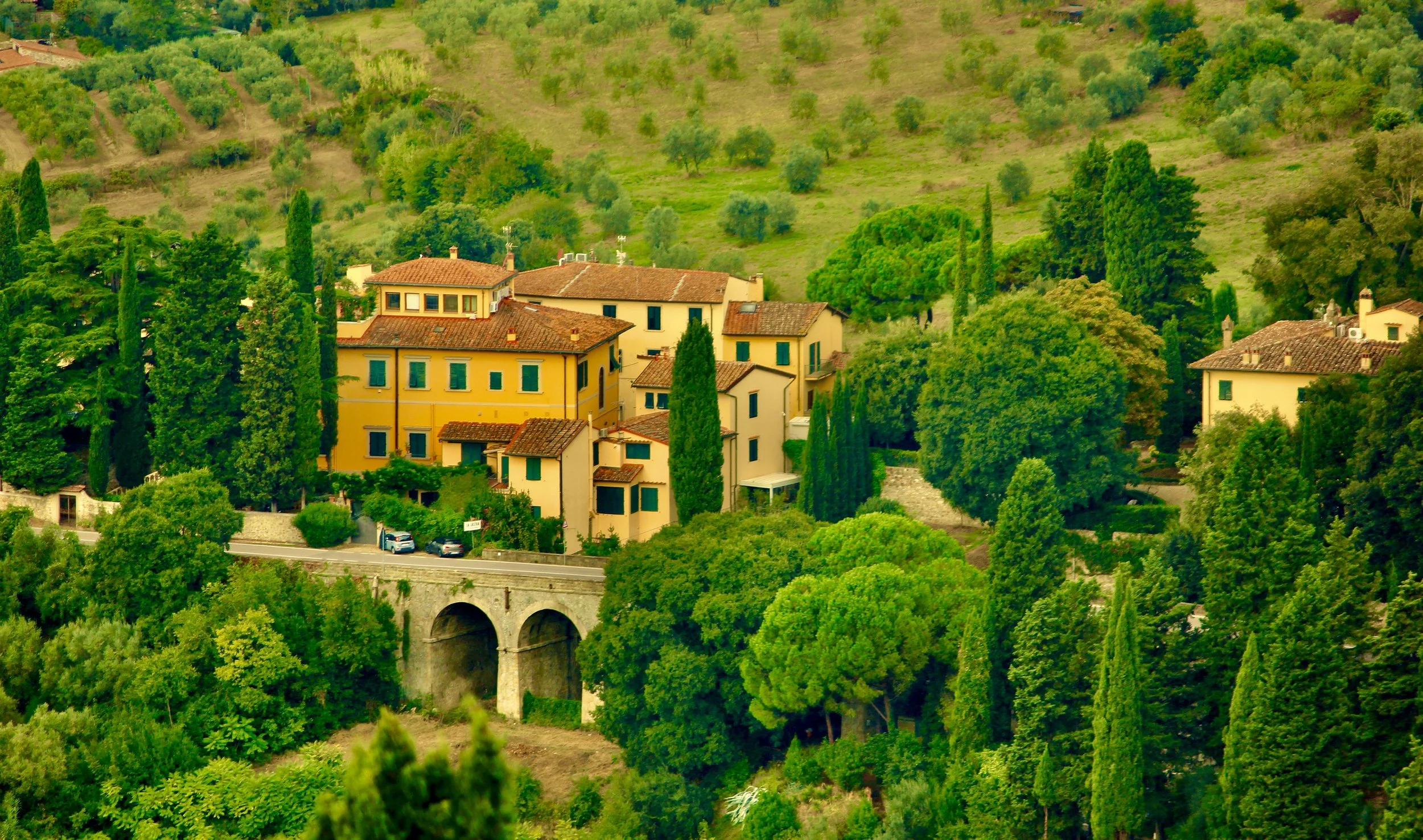 Yellow Mediterranean-style buildings with red-tiled roofs surrounded by lush green trees and hills, with a stone bridge visible below.