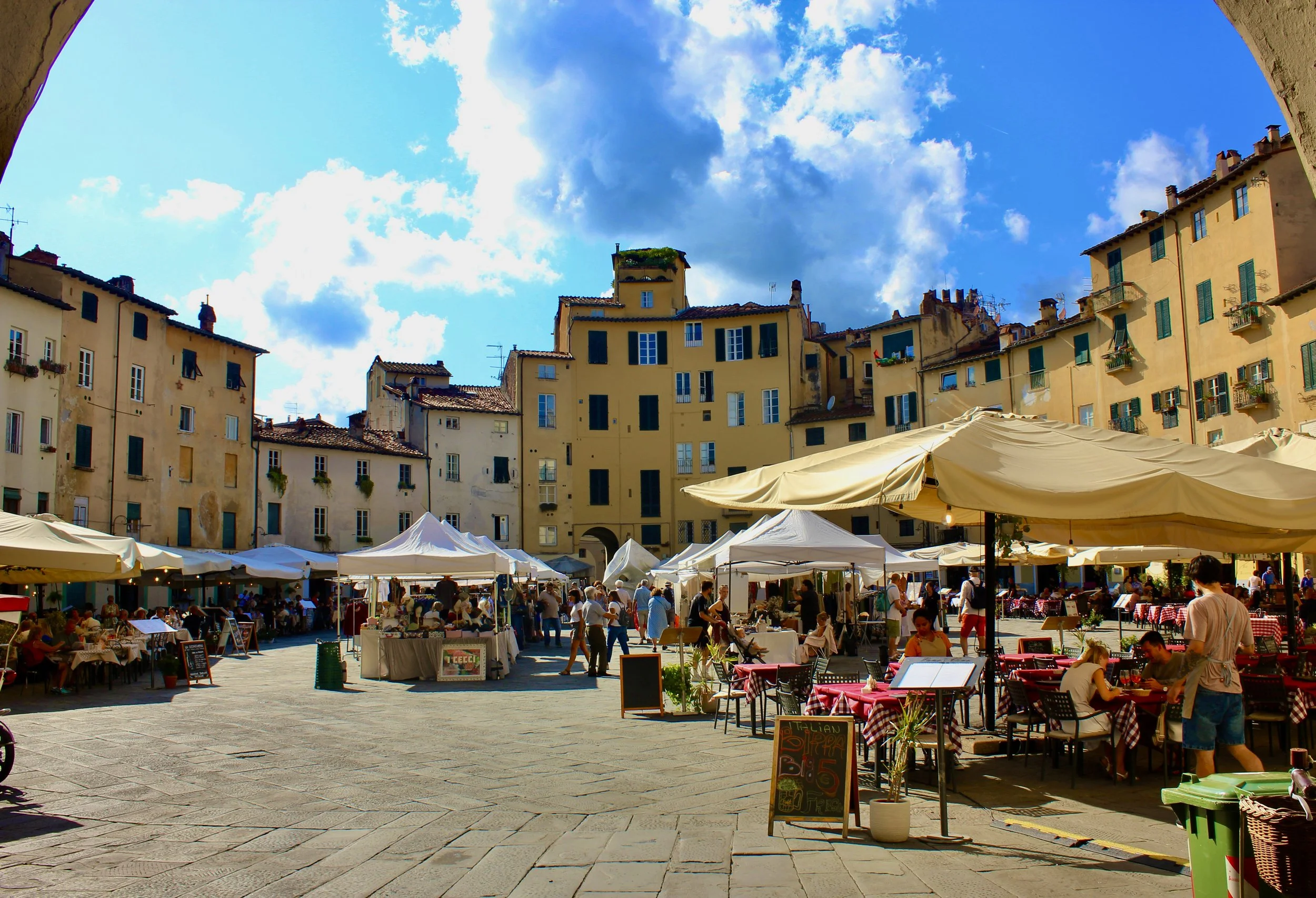 Outdoor market in a European town square with children, patrons, and vendor stalls under tents, surrounded by tall old buildings with green shutters and a partly cloudy sky.