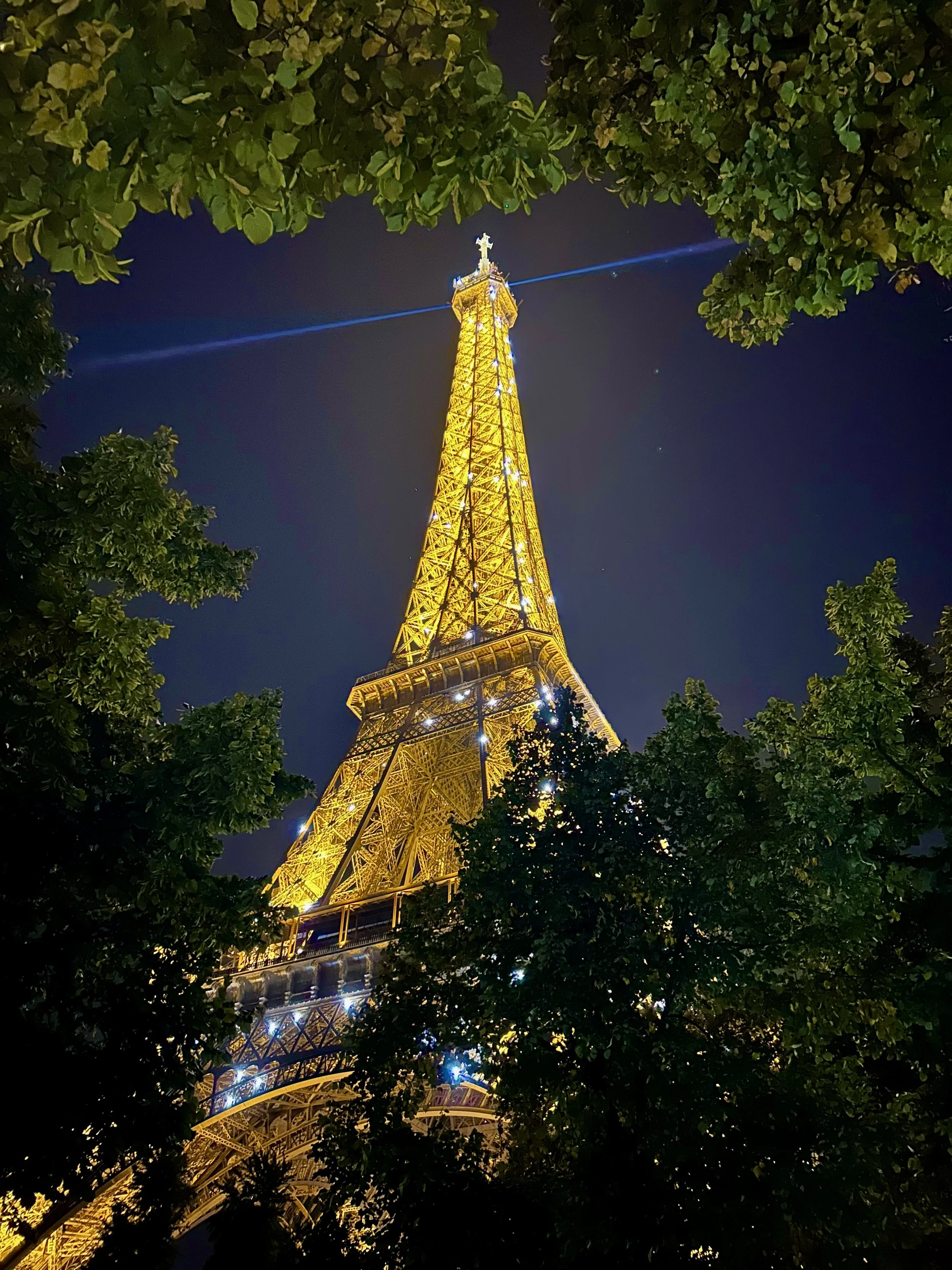 Night view of the Eiffel Tower illuminated in yellow lights, framed by green trees, with a dark sky in the background.