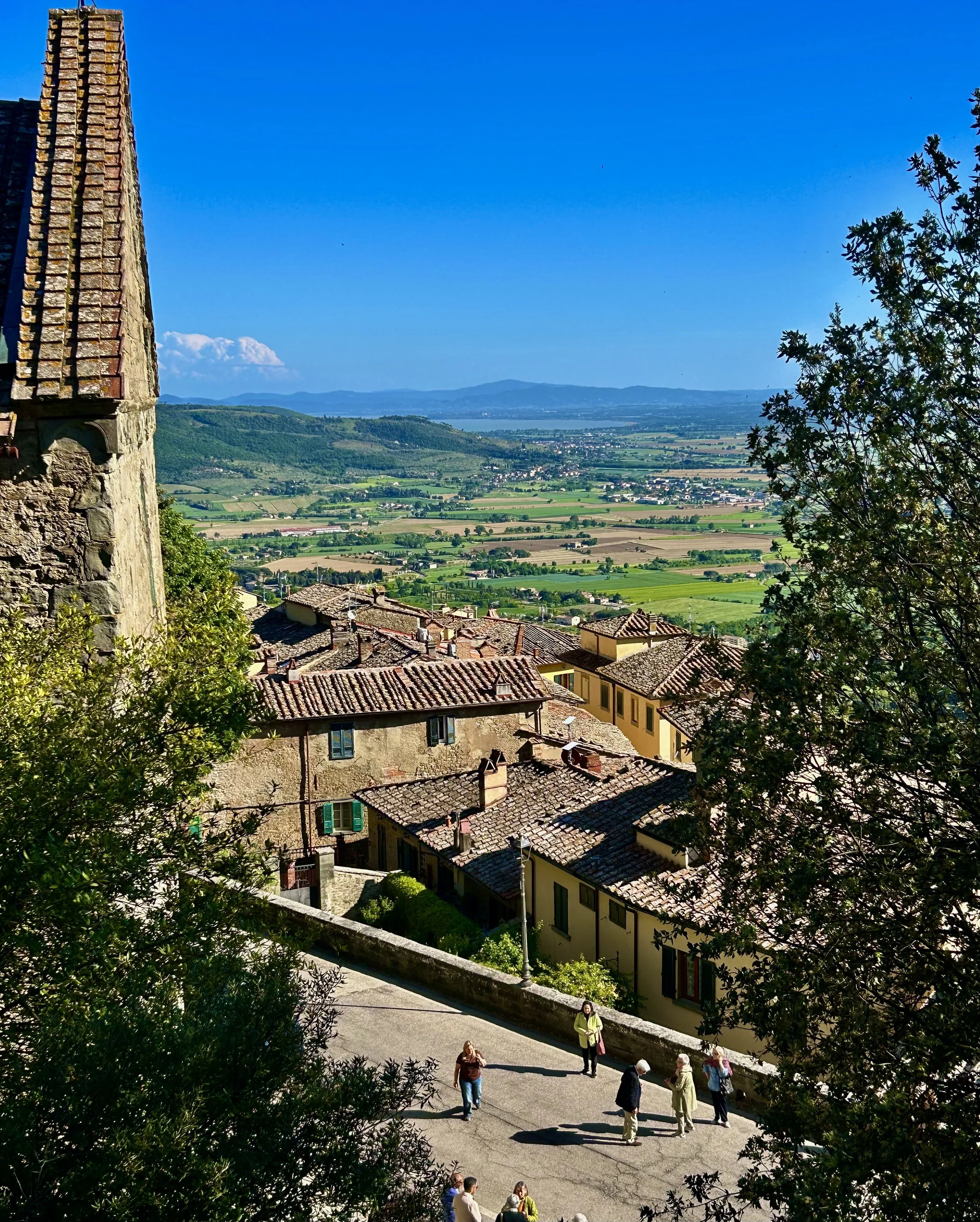 A scenic view of a small European town with tiled roofs, surrounded by lush green fields and rolling hills under a clear blue sky. A group of people is walking along a pathway in the foreground, with a historic stone building partially visible on the left.