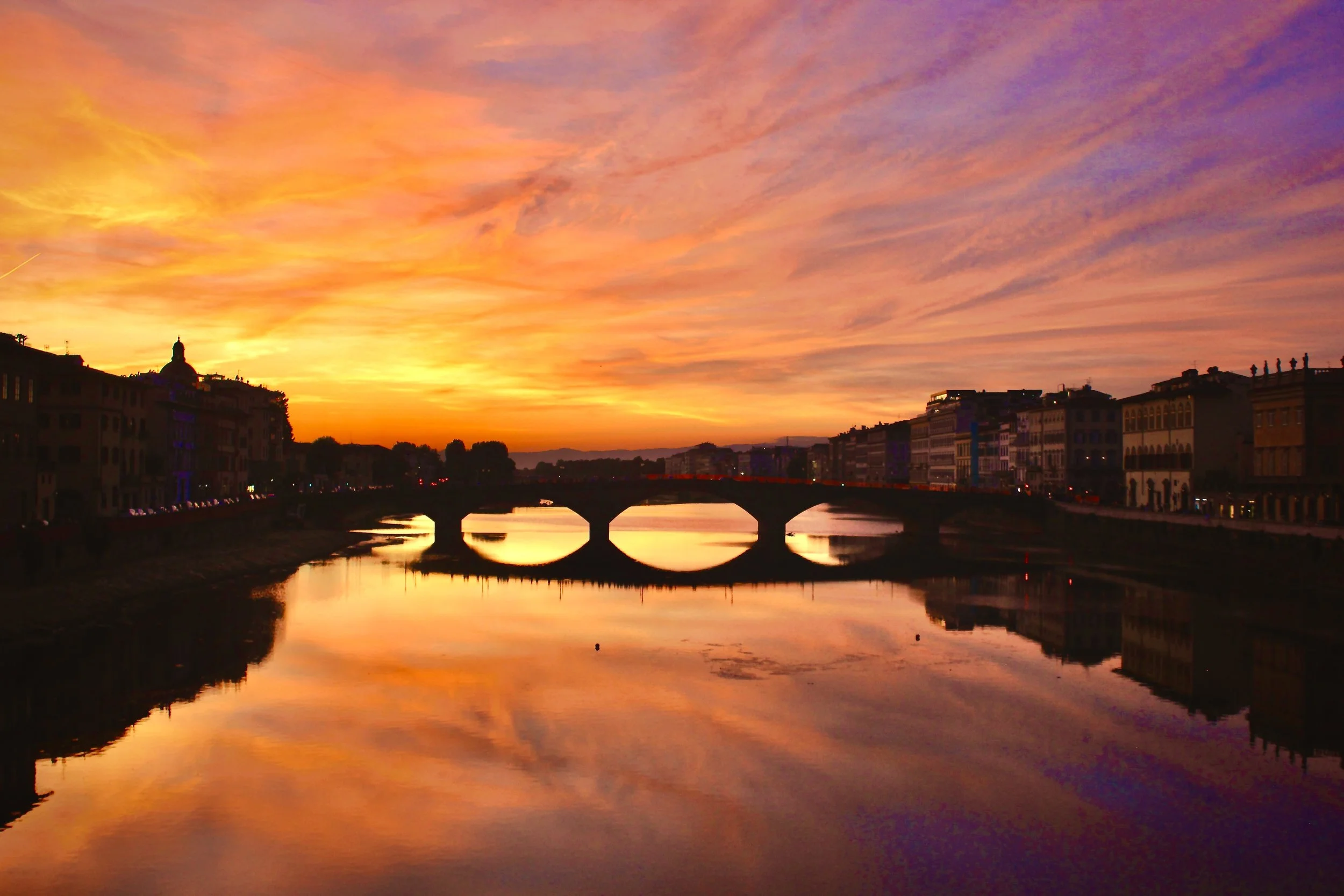 A vibrant sunset over a city skyline with colorful pink, orange, and purple clouds reflecting on a river, with buildings and a bridge in the foreground.