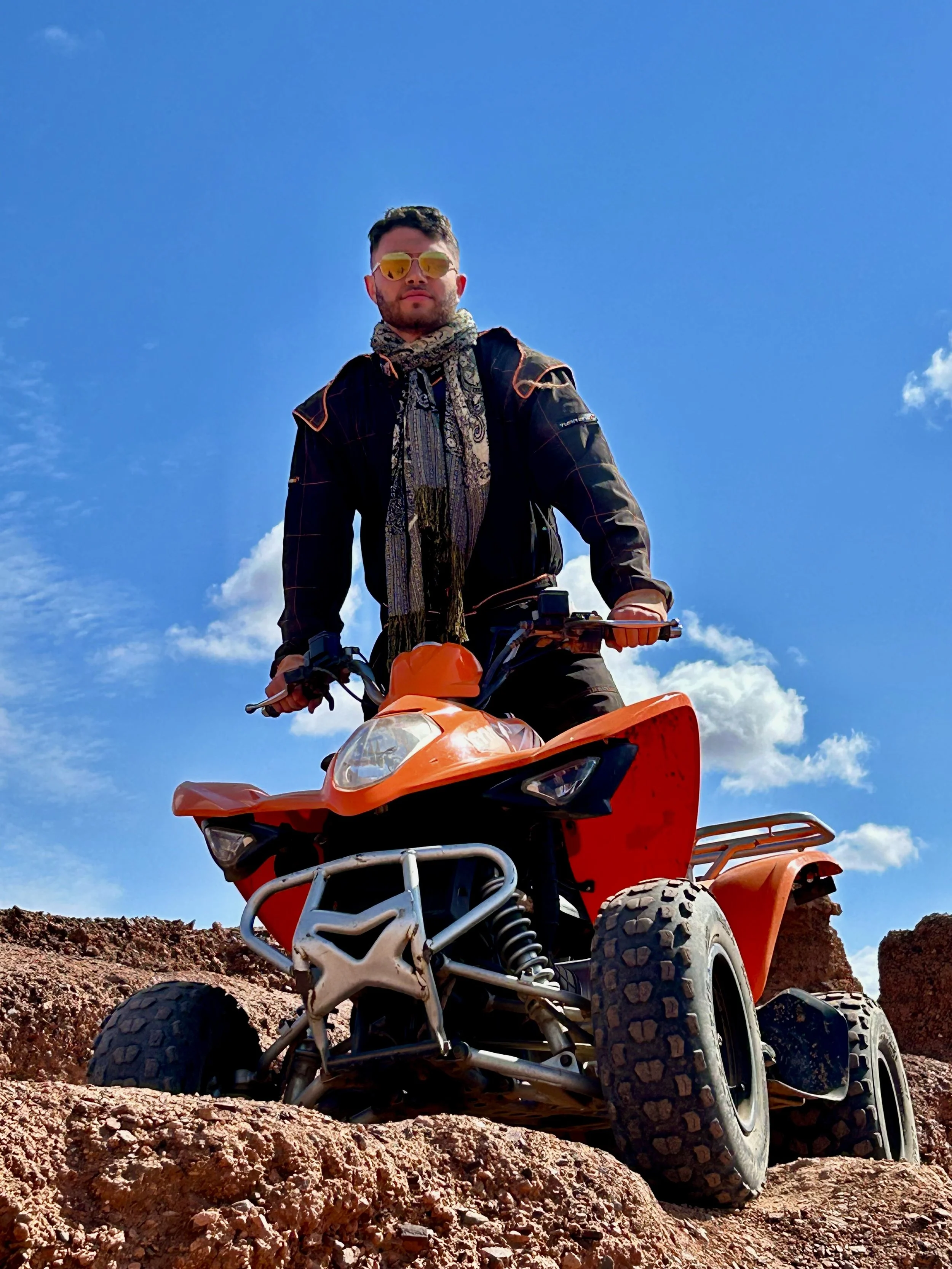 A man wearing sunglasses and a scarf rides an orange ATV on rocky terrain under a bright blue sky with some clouds.