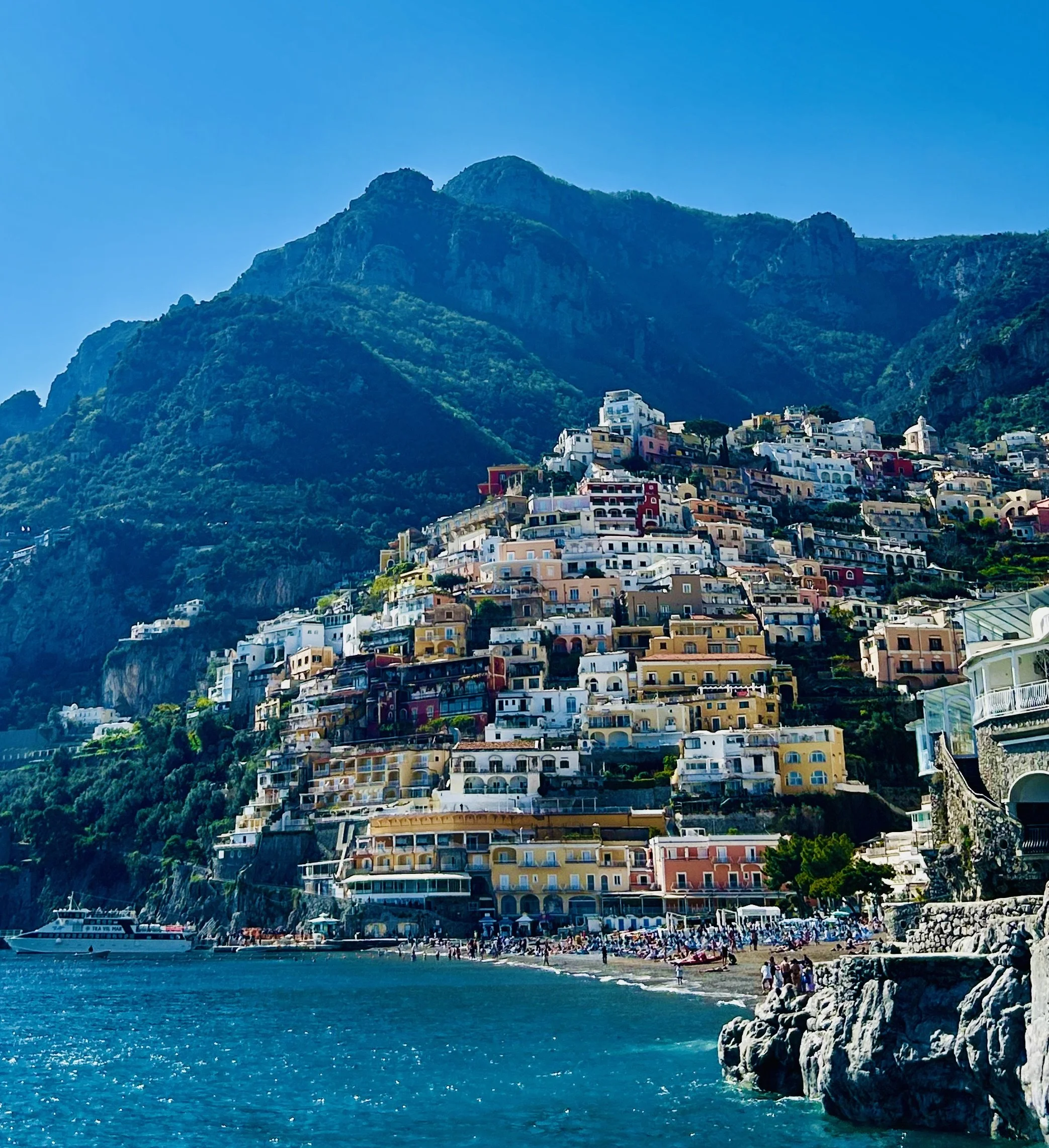 Colorful seaside town with buildings on a hillside, a sandy beach with people, and a large yacht in the water, backed by green mountains under a clear blue sky.