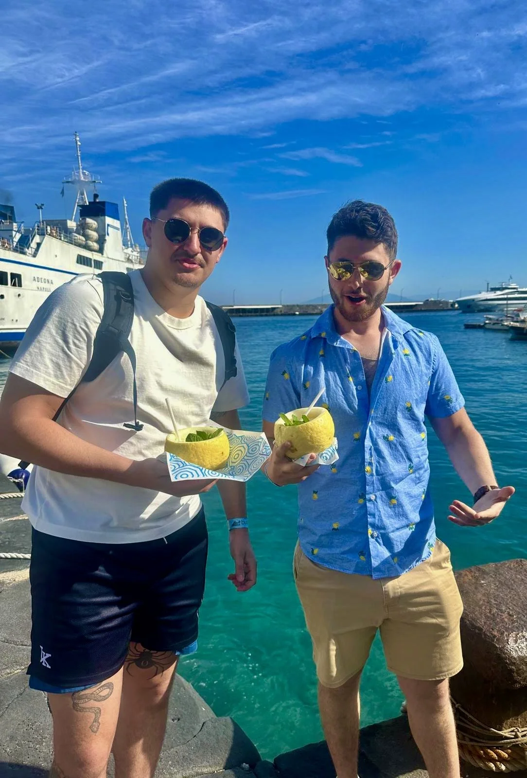 Two men standing by the water at a harbor, holding carved yellow melons filled with food and drinks, with boats and yachts in the background on a sunny day.