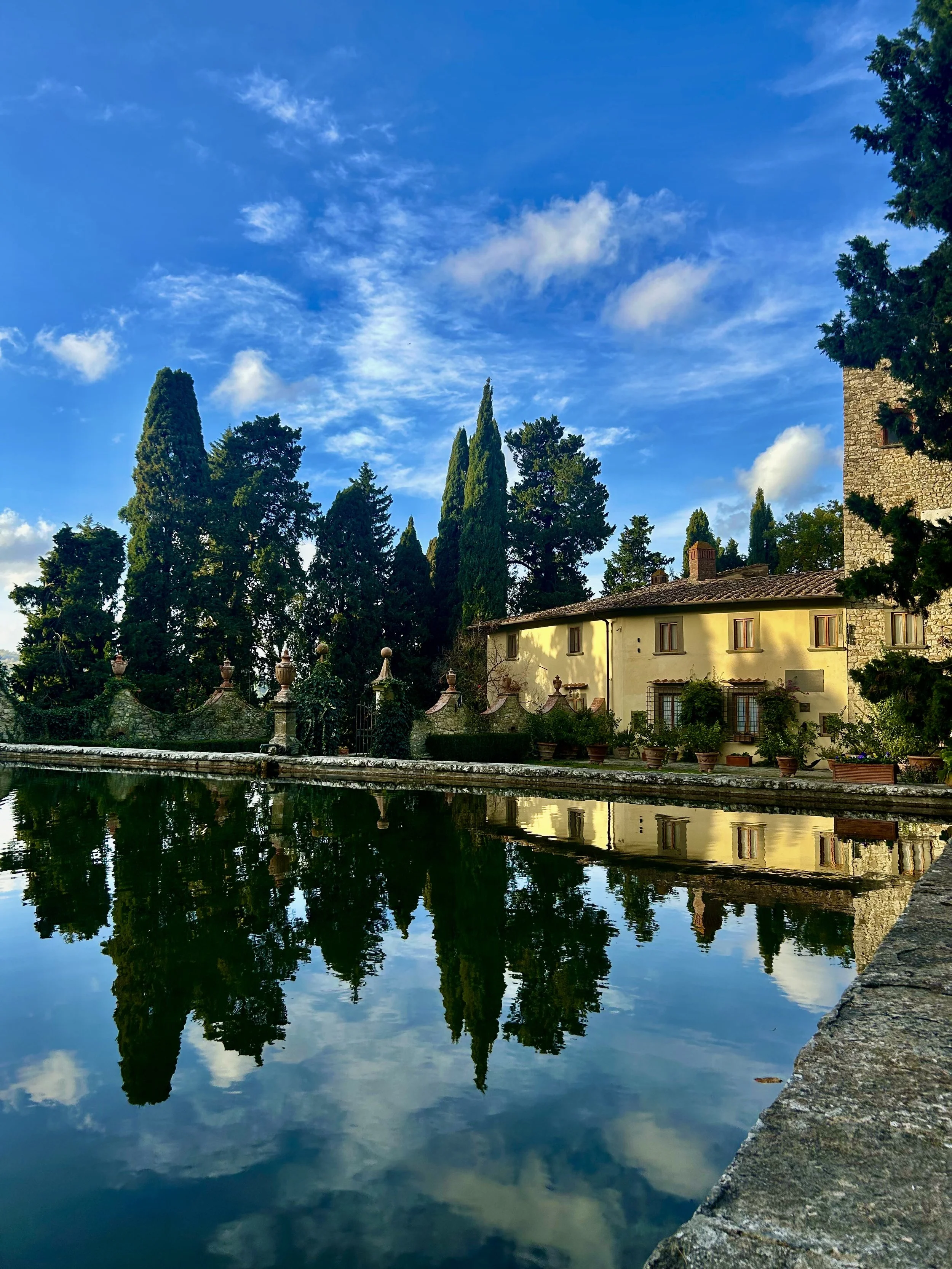 Reflection of trees, a stone building, and a blue sky with clouds in a calm body of water, with a stone edge in the foreground.