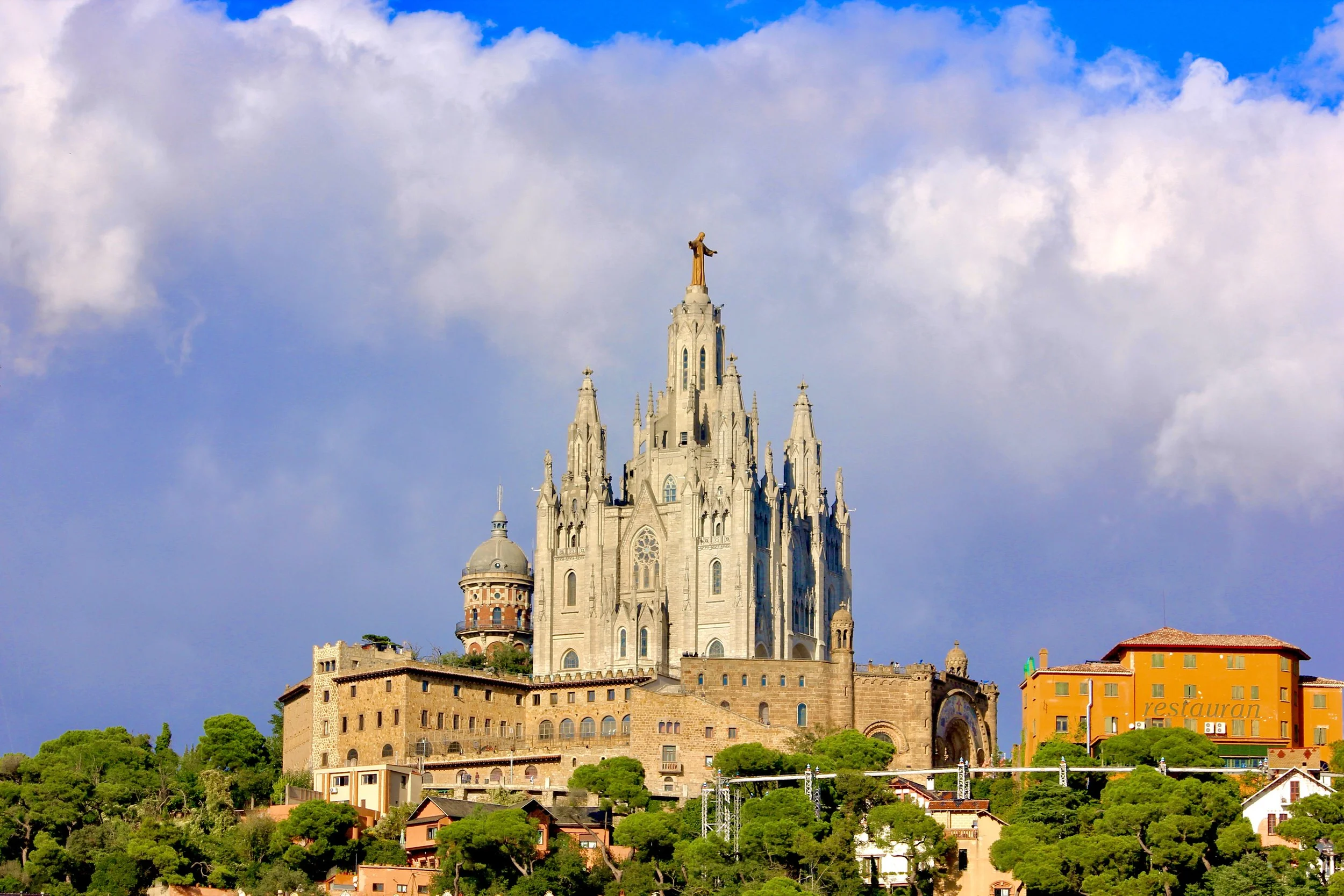 Castle with a tall spire topped with a golden statue of Christ, set against a partly cloudy sky, with greenery and buildings at the base.