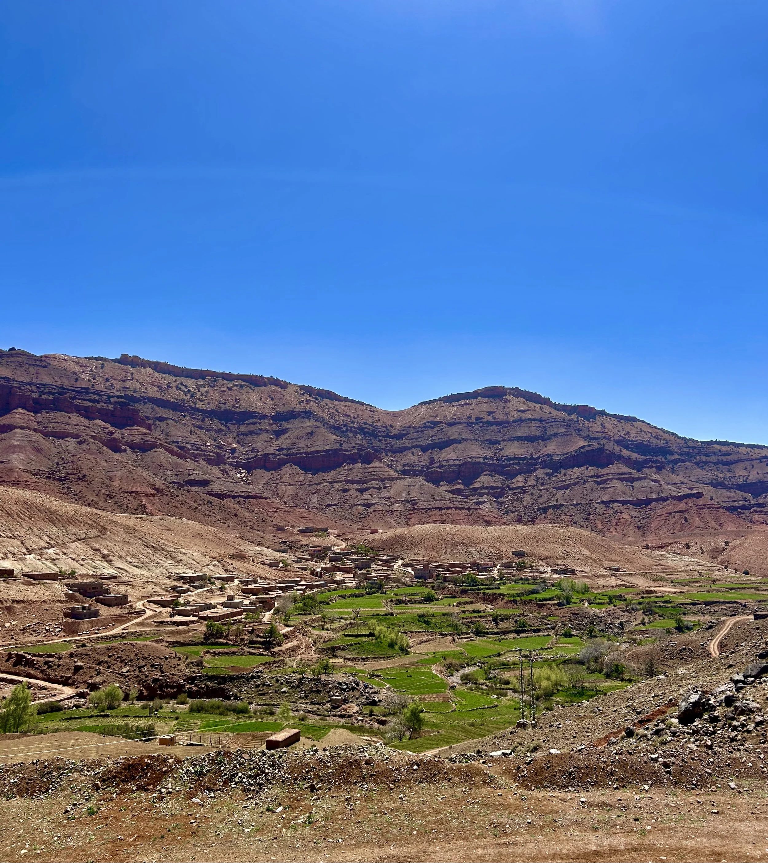 A scenic landscape showing a rural village with small houses and lush green fields surrounded by rugged, brown, rocky mountains under a clear, blue sky.
