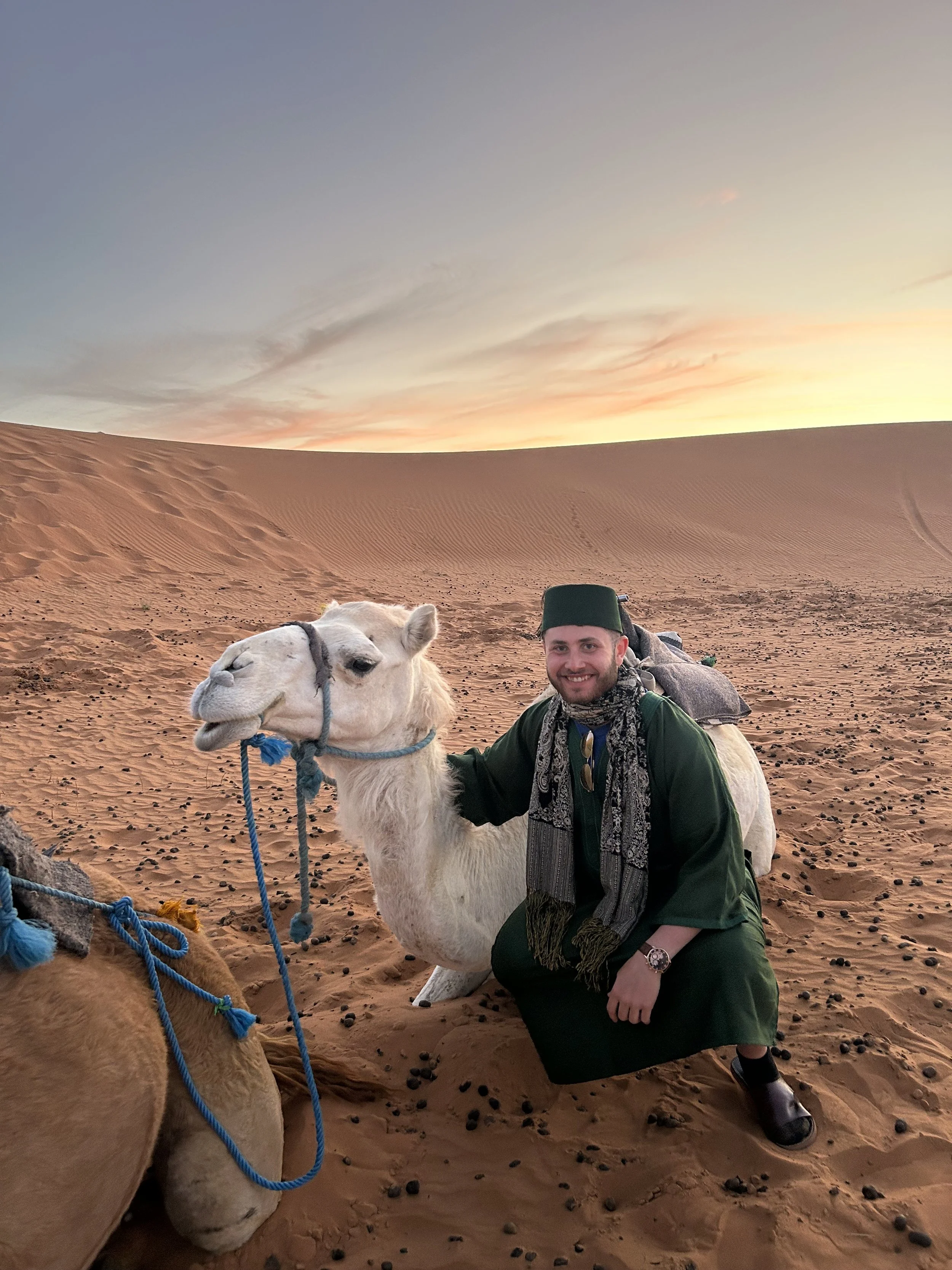 A man in desert attire crouching next to a white camel with a blue rope halter in a sandy desert landscape at sunset.