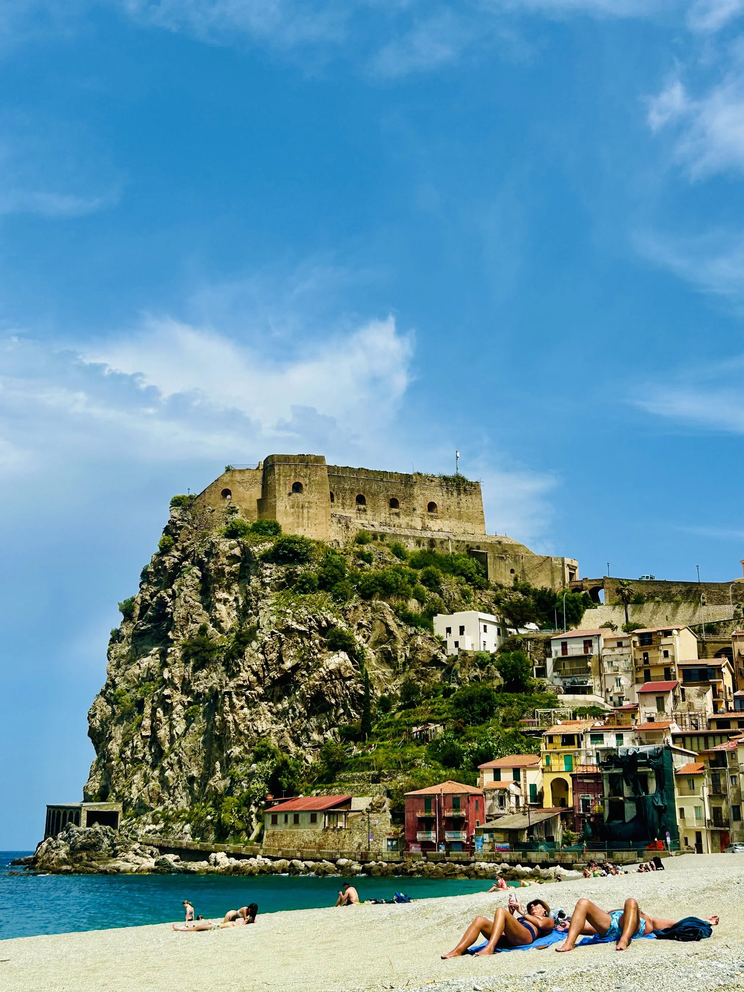 Beach scene with people sunbathing and swimming near colorful houses on a hillside under a blue sky, with a fortress on a rocky hill in the background.