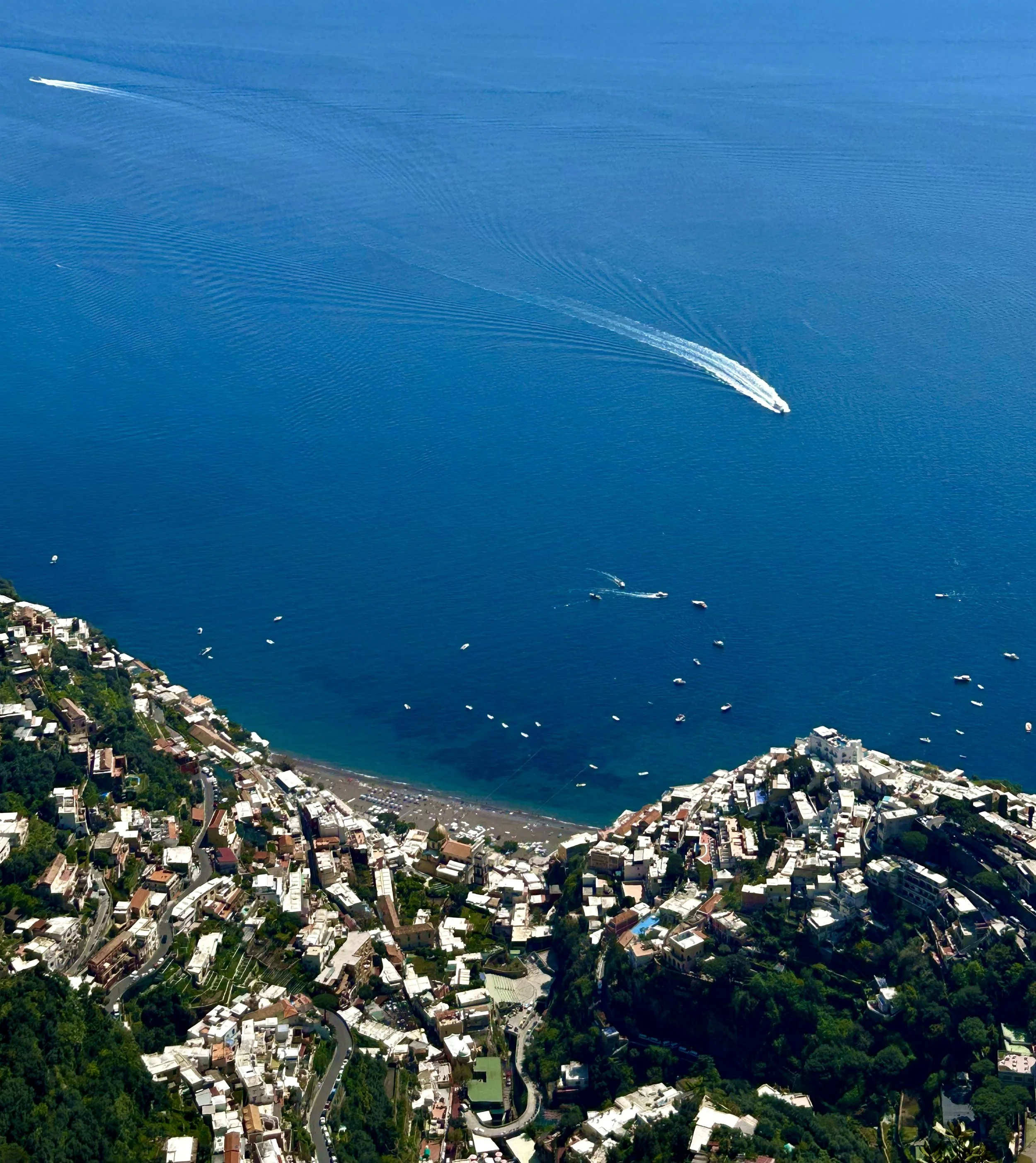 Aerial view of a coastal town with buildings along the shoreline, winding roads through lush green hills, and boats in the water. A large cruise ship is sailing in the blue ocean, leaving a wake behind.