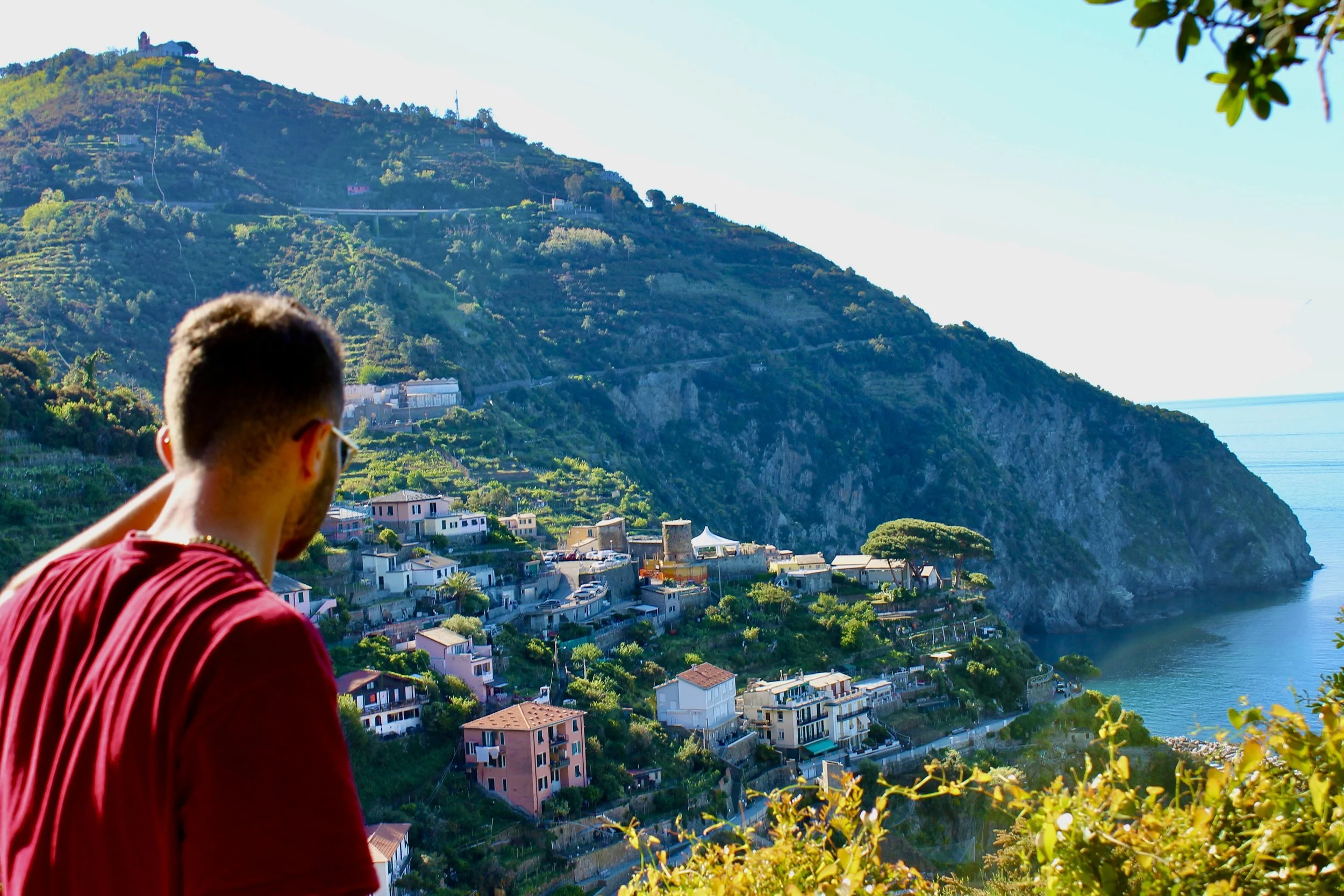 Man in red shirt looking over coastal hillside town with houses, greenery, and a mountain in the background.