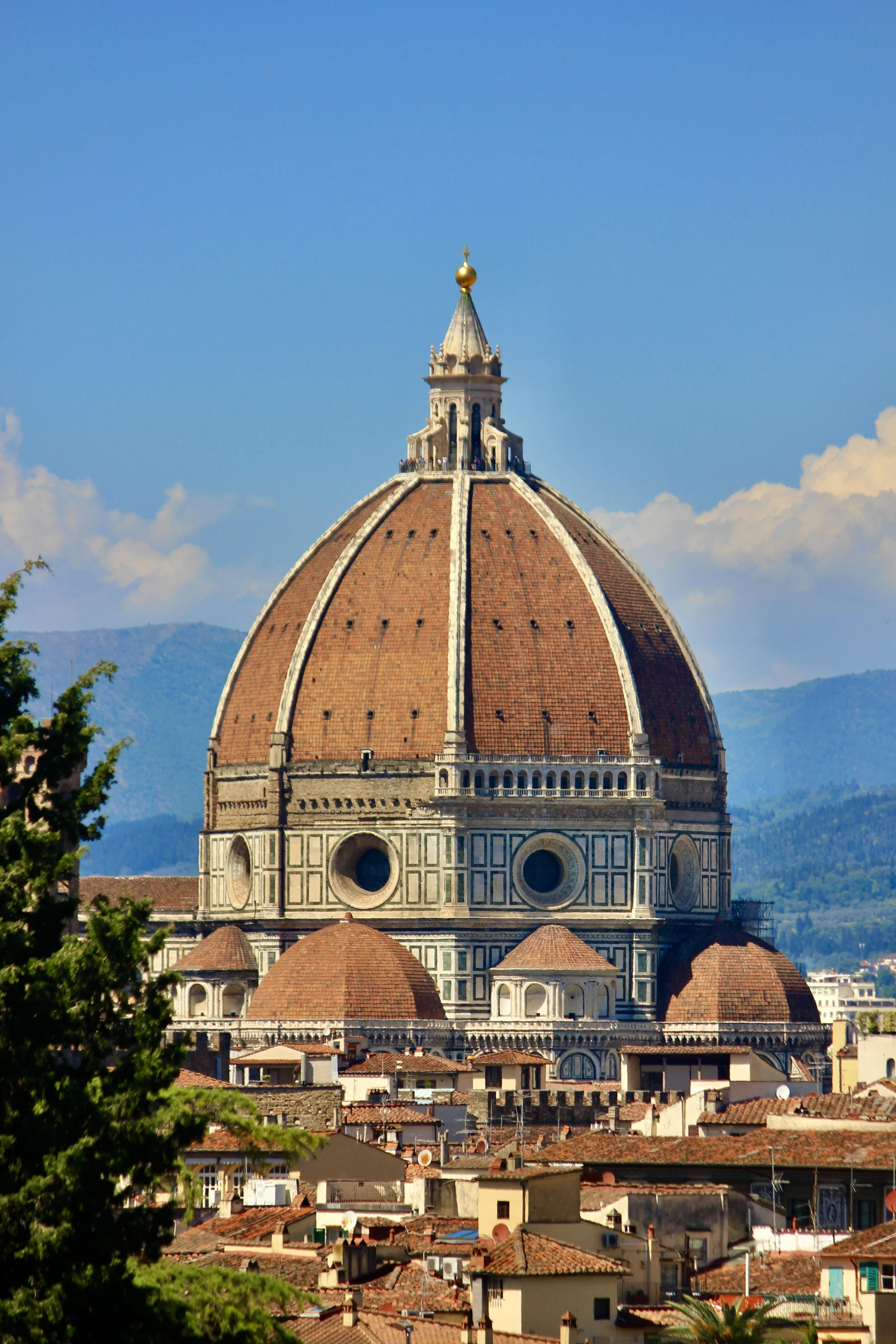 The Florence Cathedral, also known as Cattedrale di Santa Maria del Fiore, with a large red-tiled dome topped by a small golden sphere and cross, set against a bright blue sky with some clouds.