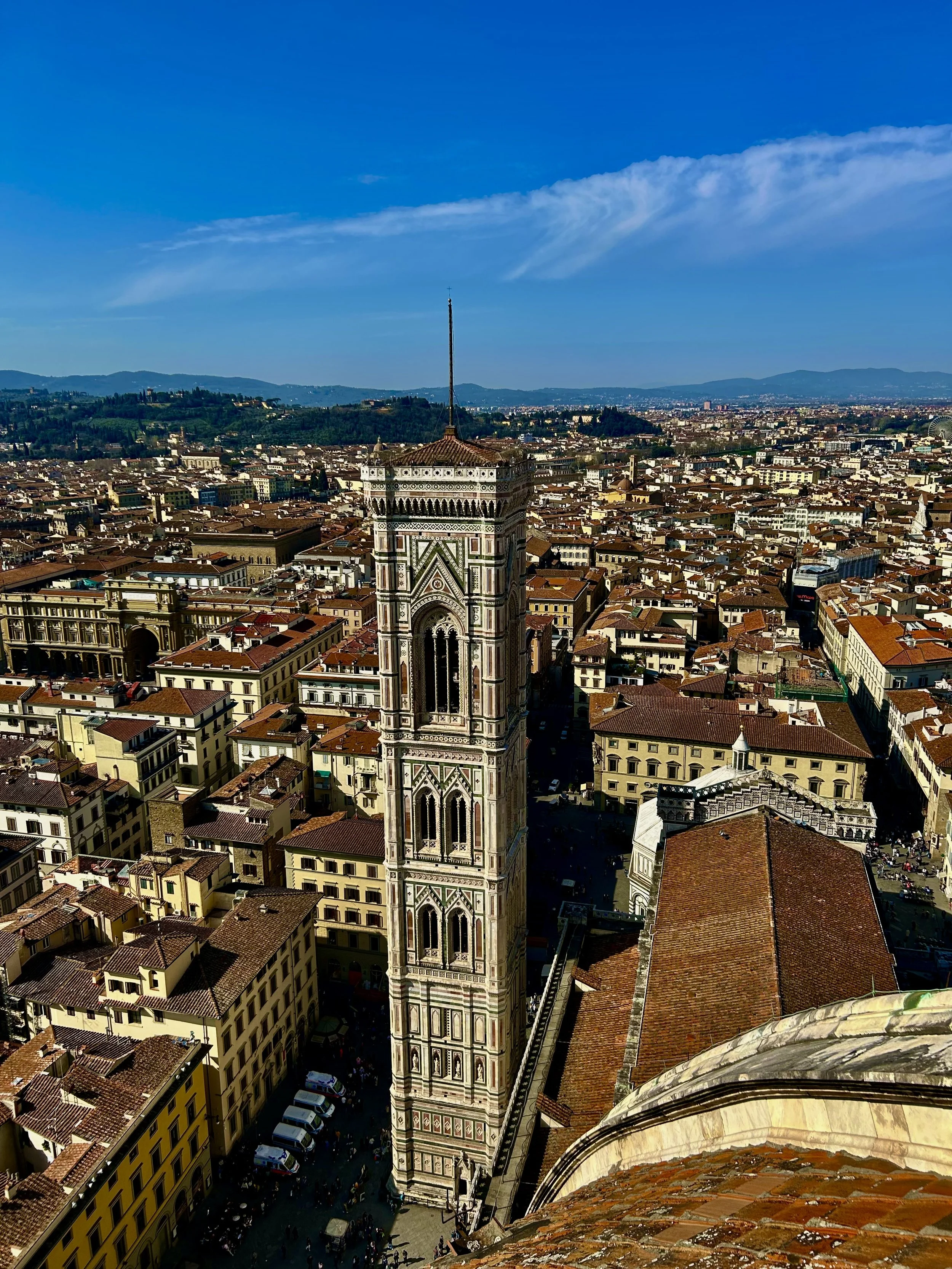 Aerial view of a historic city with a tall, intricately decorated bell tower in the foreground. The cityscape includes many terracotta rooftops and a mix of old and modern buildings, with rolling hills and mountains in the background under a clear bl