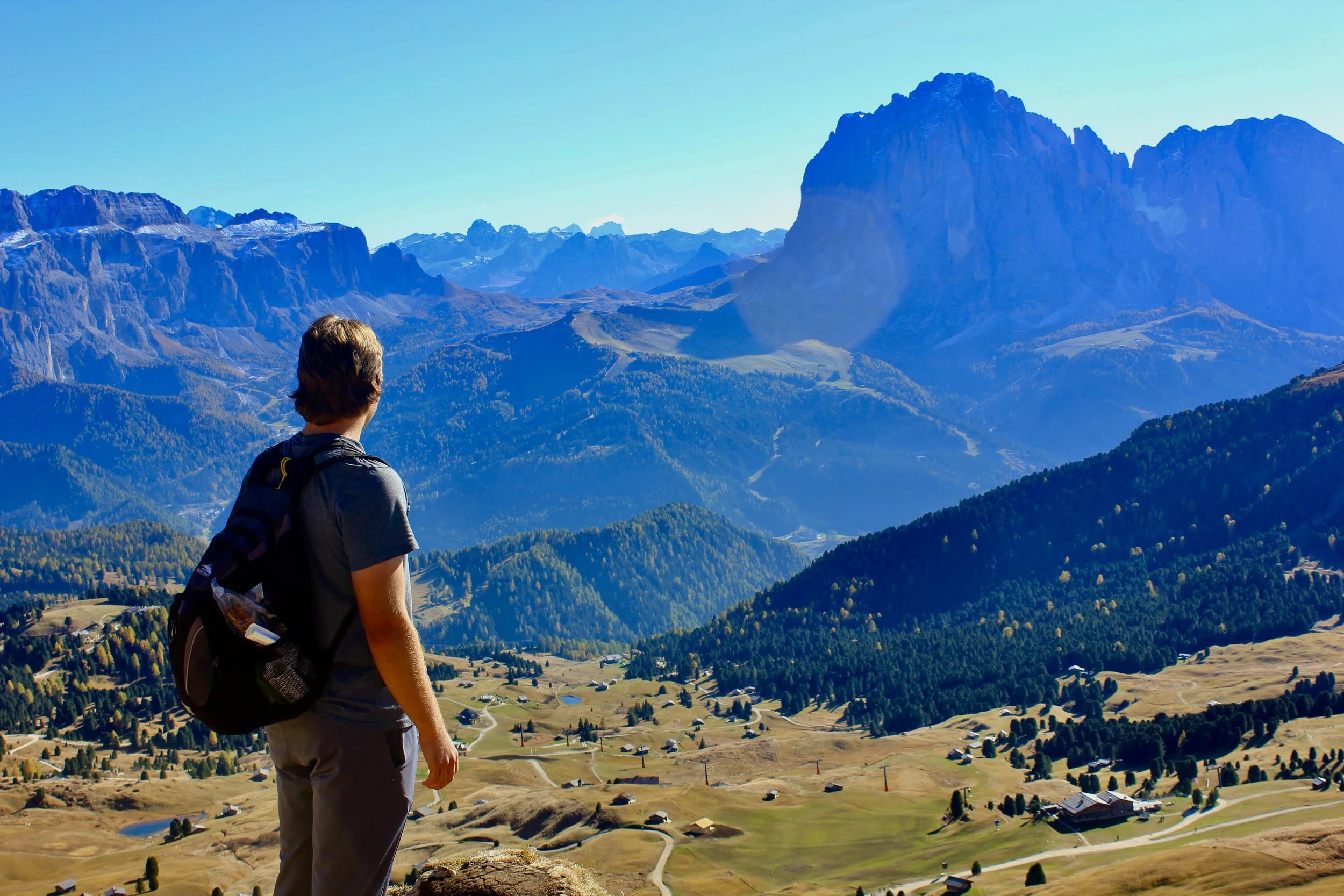 A person standing on a hilltop overlooking a valley with small houses, surrounded by green and brown fields, with mountains in the distance under a clear blue sky.