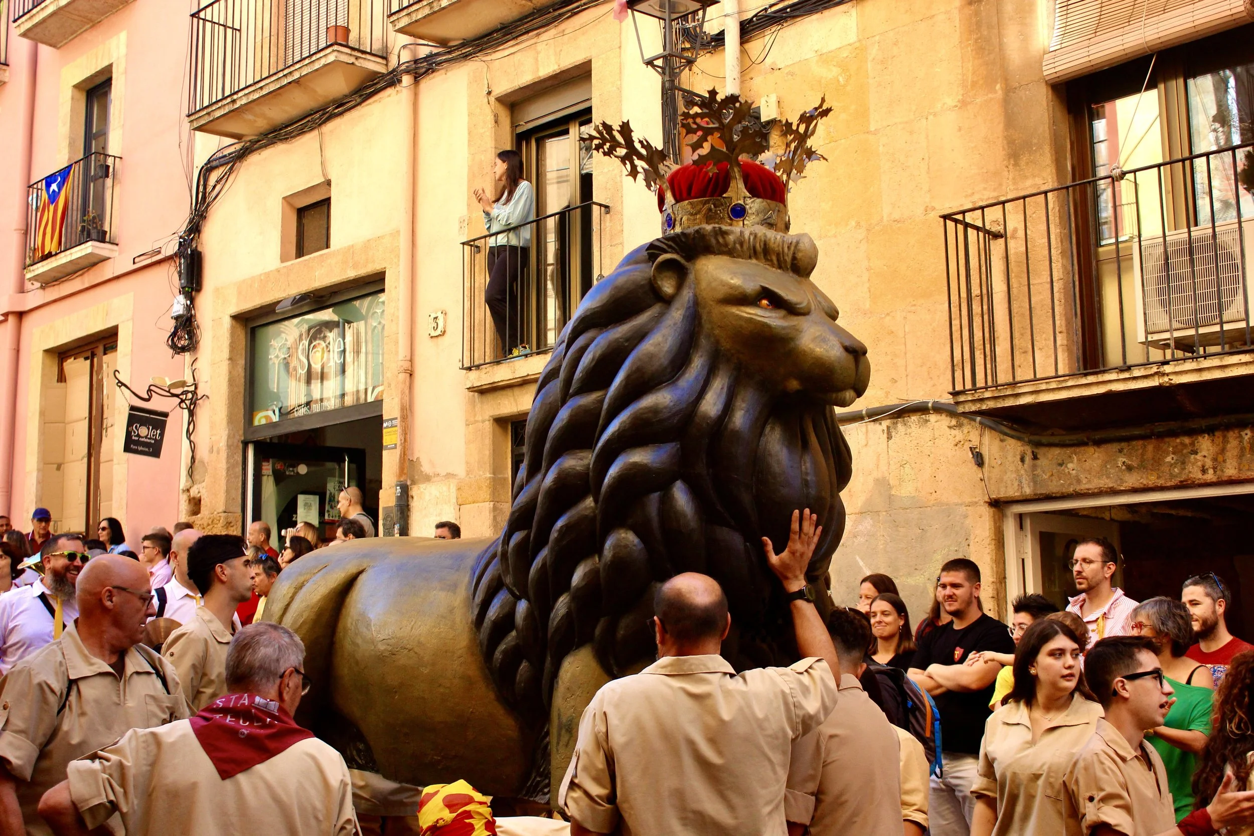A crowd of people gathers around a large bronze lion statue with a crown and decorative elements on its head during a festival on a city street. Some individuals are standing on balconies of the buildings in the background, watching the event.