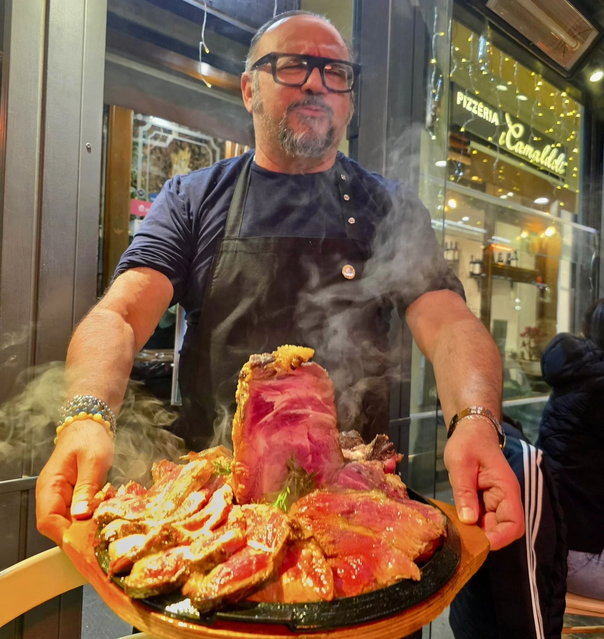 A man with glasses and a beard, wearing a black apron, stands behind a very large plate of assorted sashimi, with steam rising from it, inside a restaurant with a neon sign in the background.