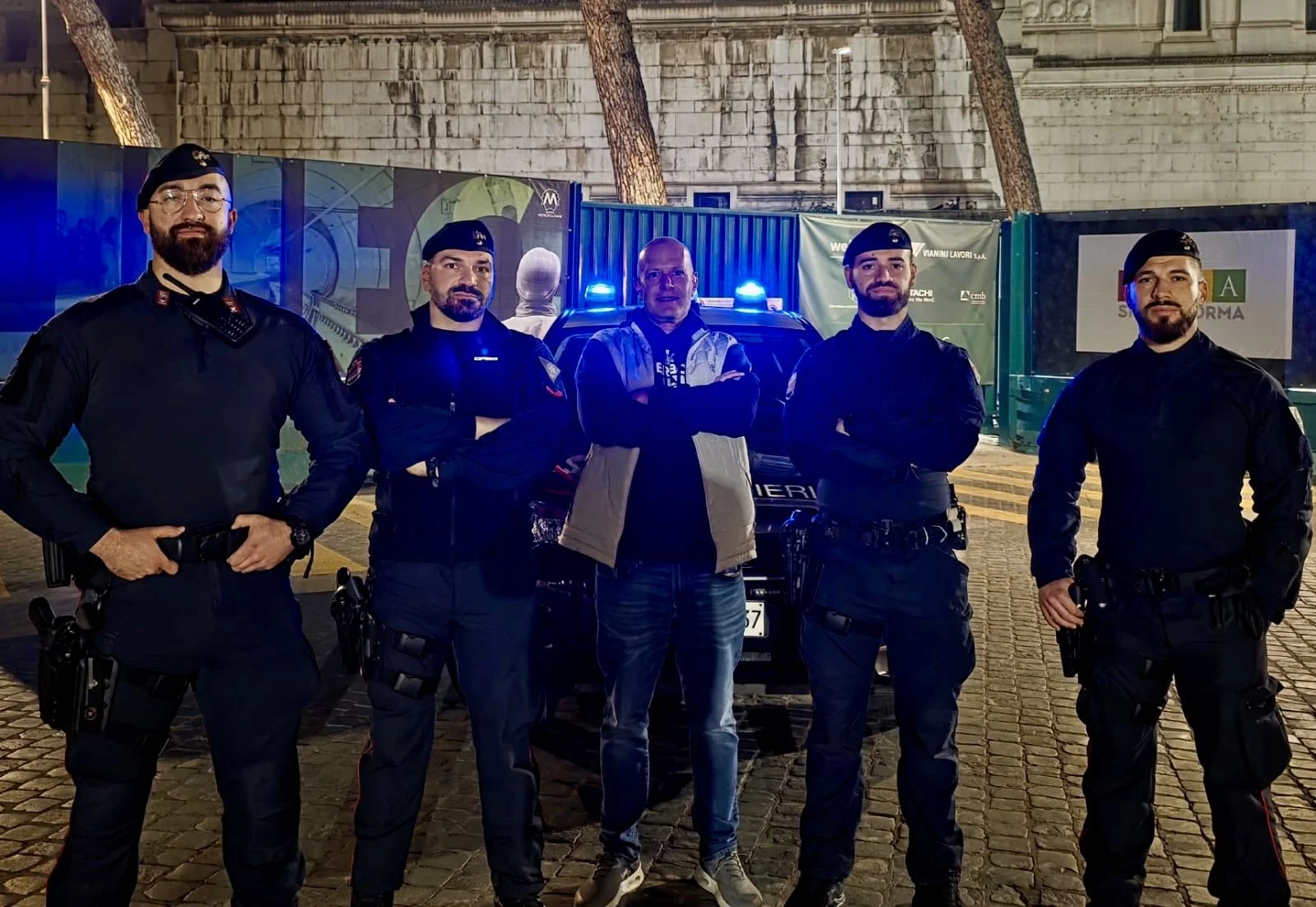 Four police officers and a civilian man standing in front of a police car with blue lights on a cobblestone street at night. The officers are wearing black uniforms with hats, and the man is dressed casually. There are trees and a stone building in t