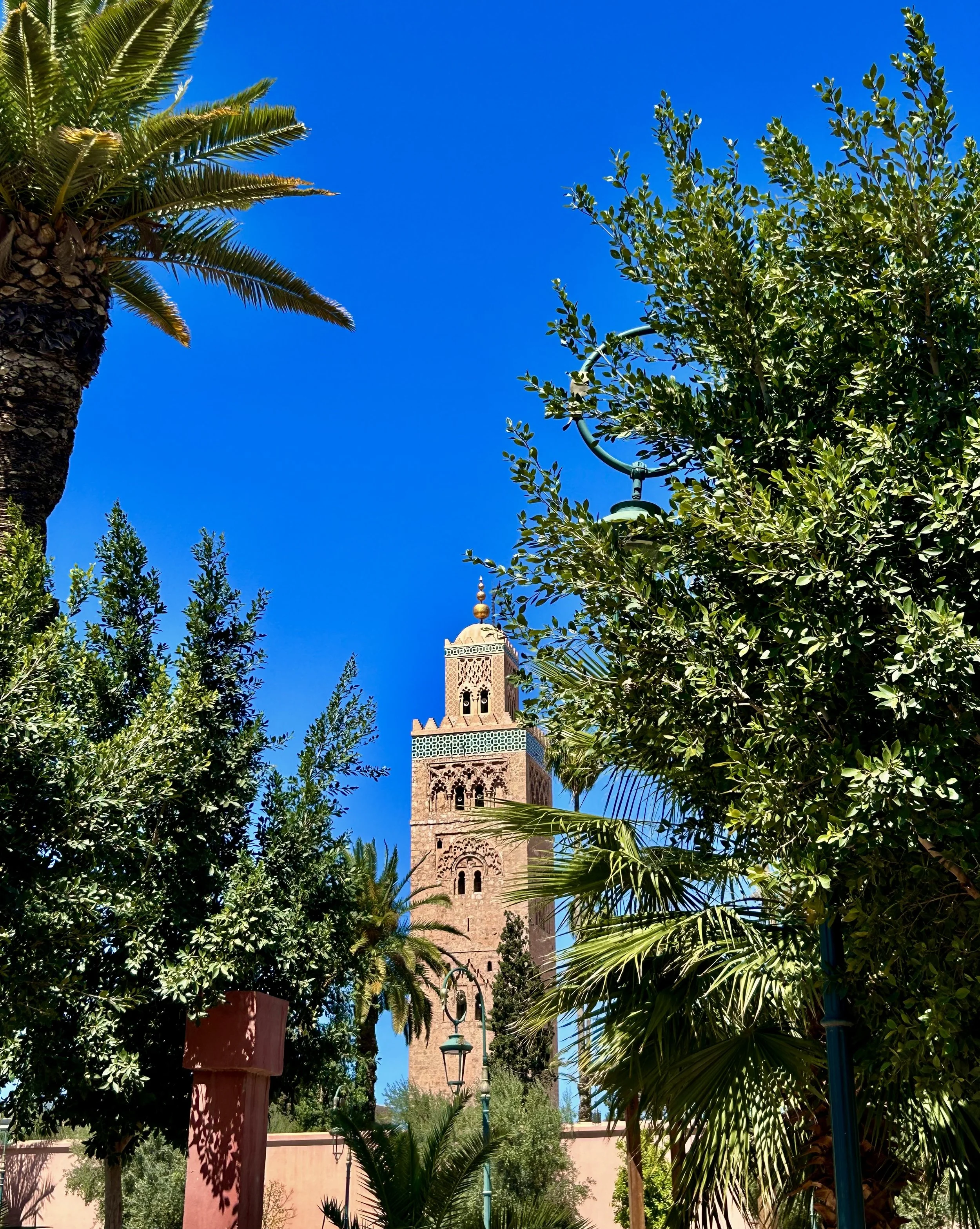 A tall, ornate tower with a golden dome is framed by lush green palm trees and foliage under a clear blue sky.