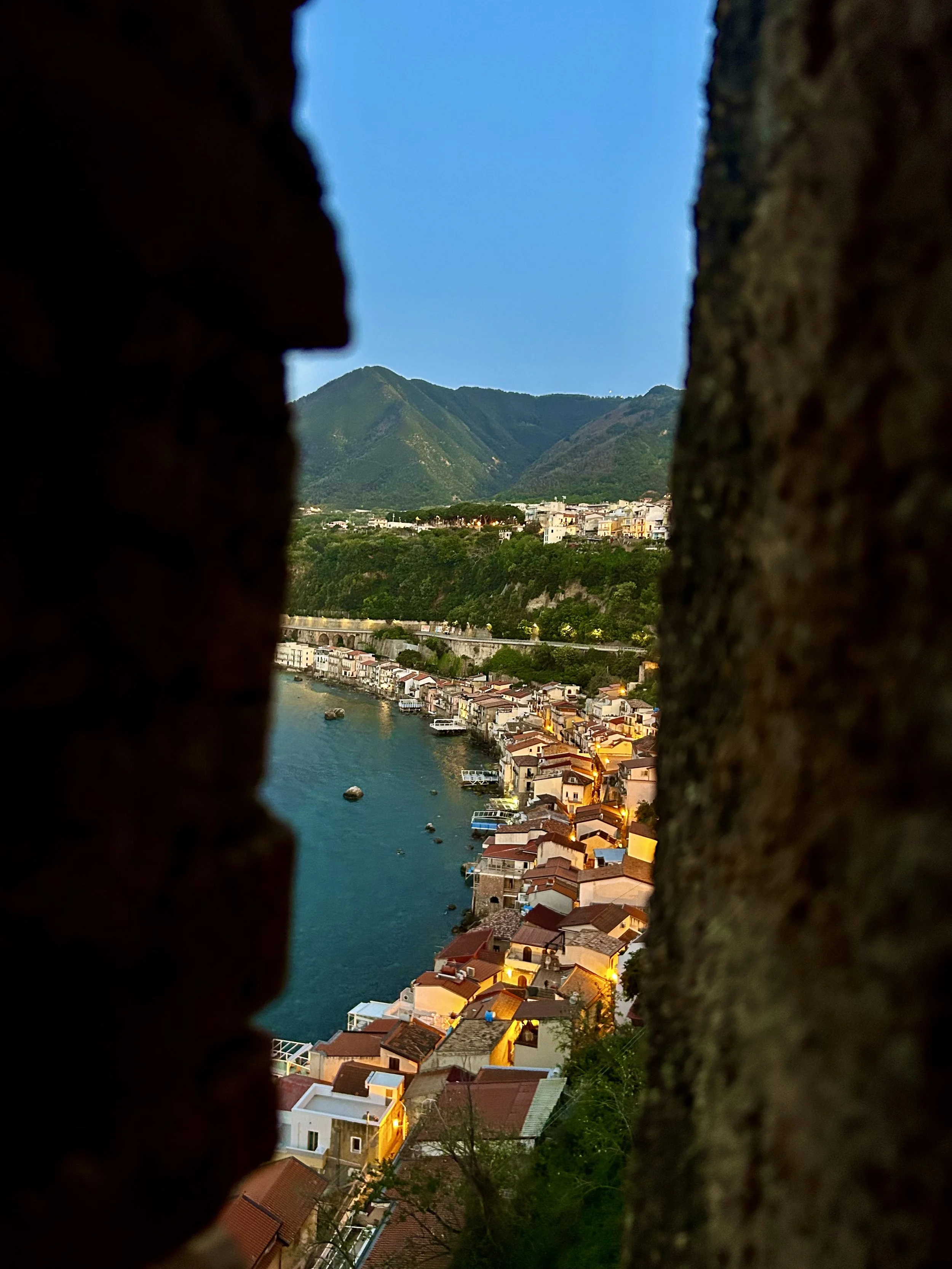 View of a coastal town with colorful houses along the water, mountains in the background, seen through a narrow opening between two brick walls during dusk.