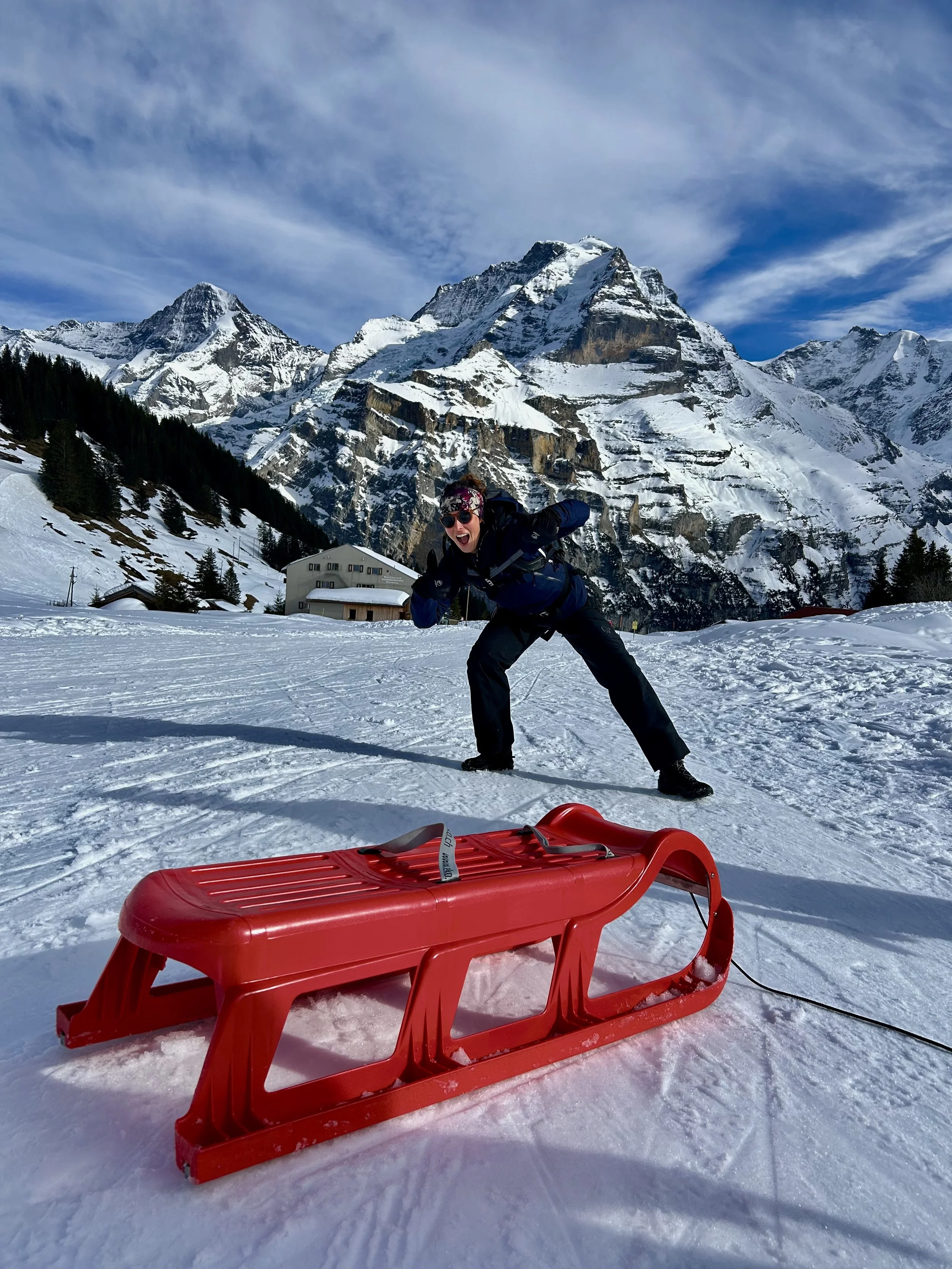 A woman in winter gear smiling and posing on snow-covered ground with snow-capped mountains and a partly cloudy sky in the background, and a red sled in the foreground.