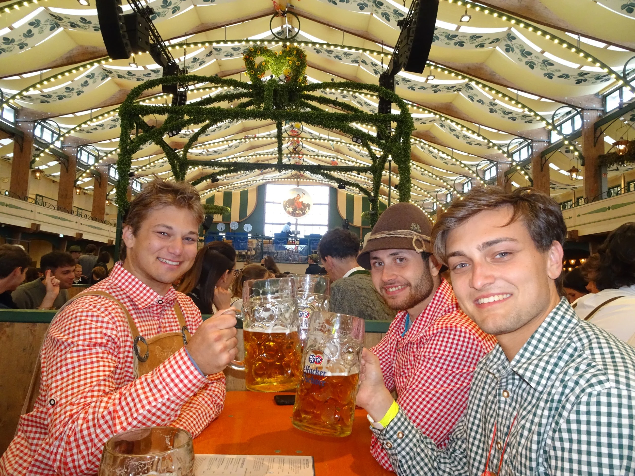 Three young men wearing traditional German/checkered shirts toasting with large beer steins in a festive indoor setting with decorative hanging heart and greenery, large roof with patterned paneling, and crowd of people in the background.