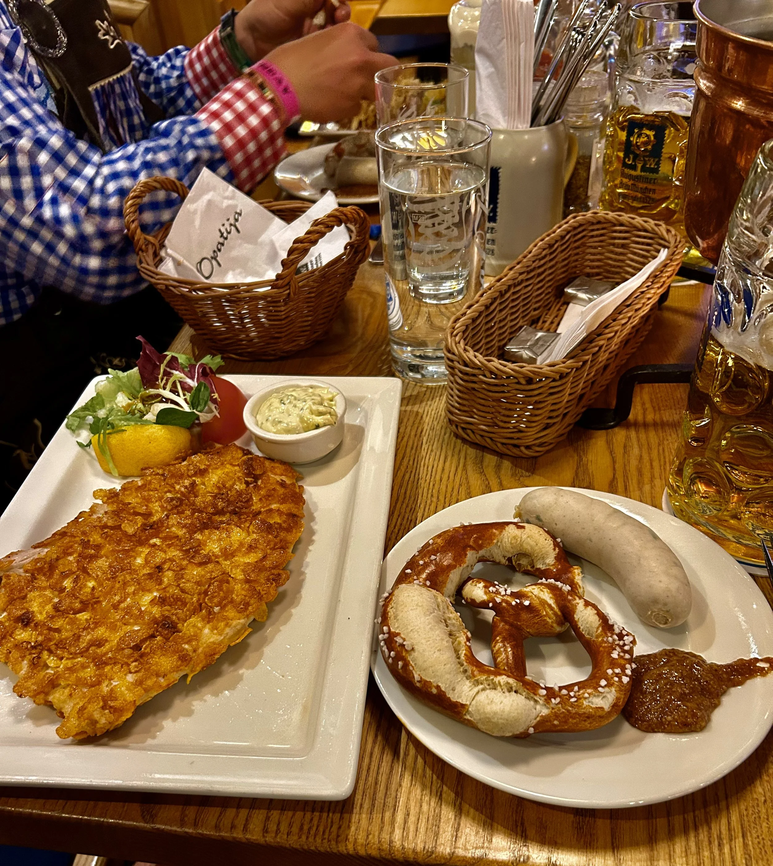 A table with assorted foods including a large fried fish fillet, salad, bread rolls, pretzels, and condiments, with beverages such as water and beer in the background, and guests in checkered shirts dining indoors.