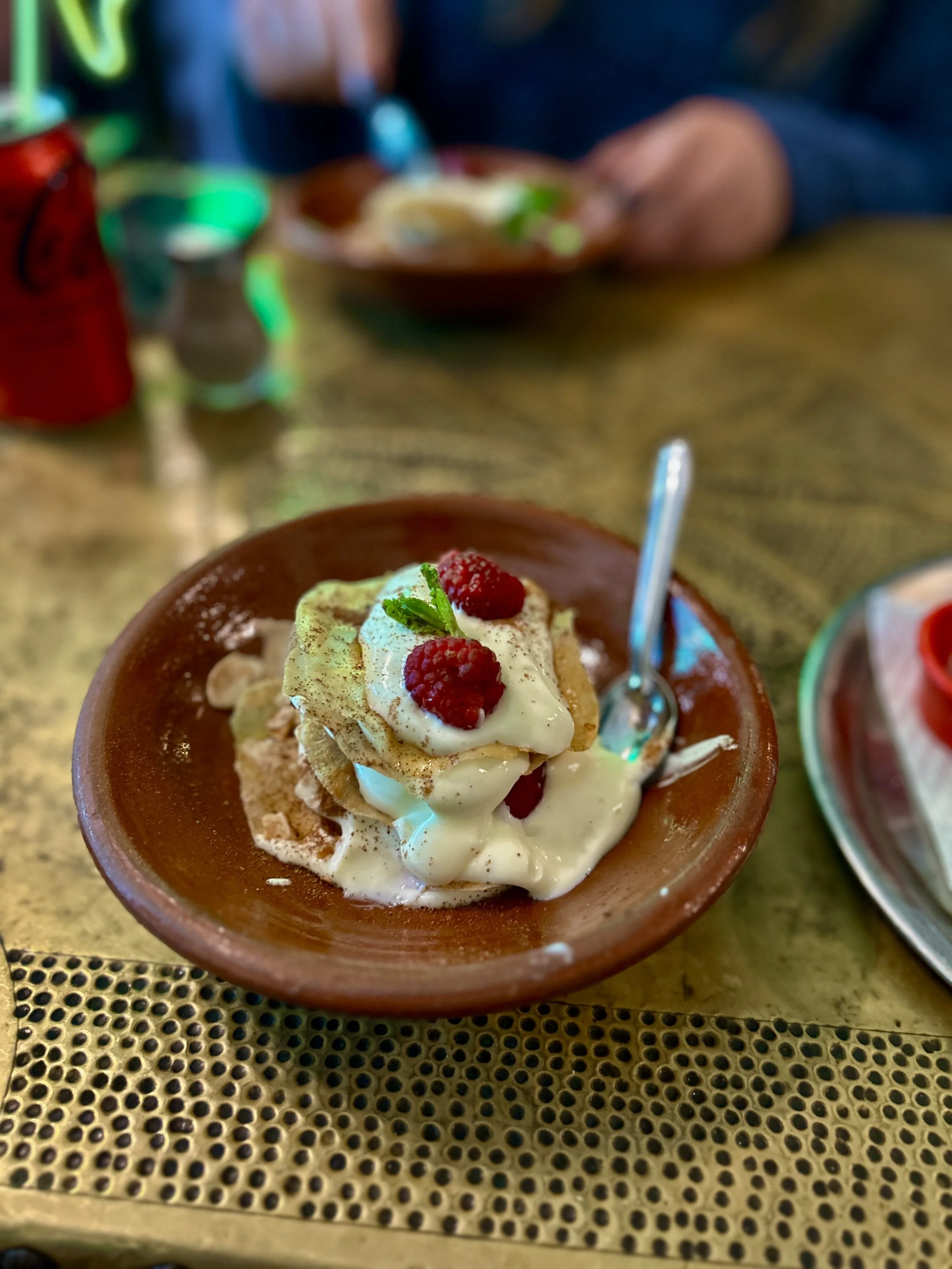 A bowl of ice cream topped with raspberries, a sprig of mint, and a biscuit shard, served in a small brown dish on a textured yellow surface.