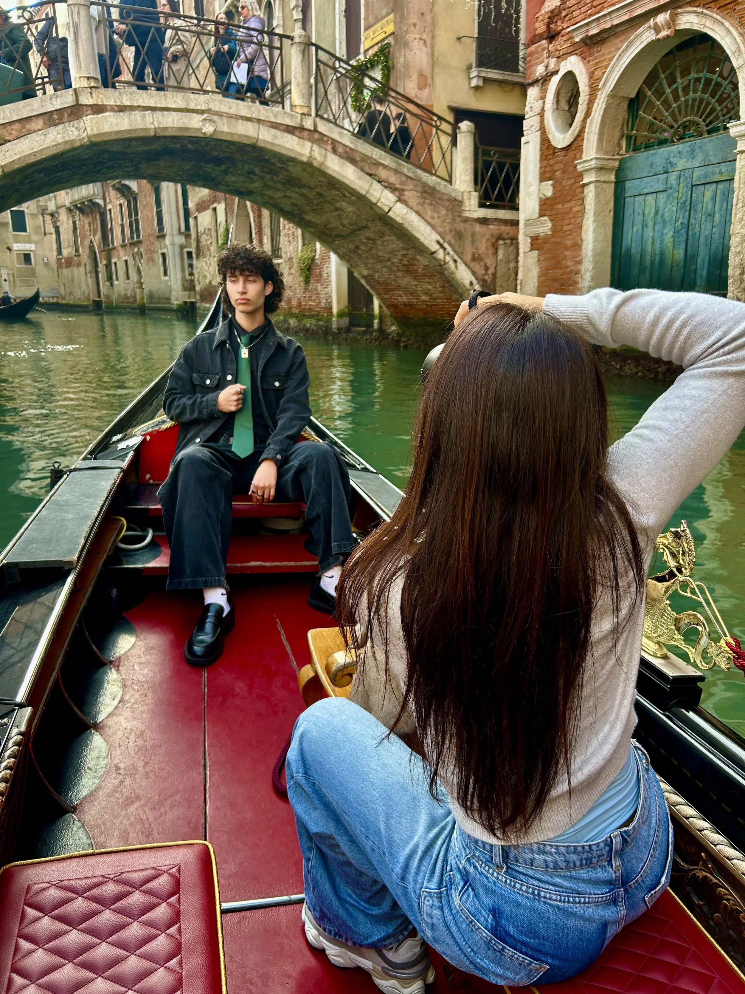 A person with long brown hair sitting on a gondola in Venice, Italy, taking a photo of a young man with curly dark hair, wearing a black jacket and green tie, sitting on the gondola. The canal and stone bridge are visible in the background.