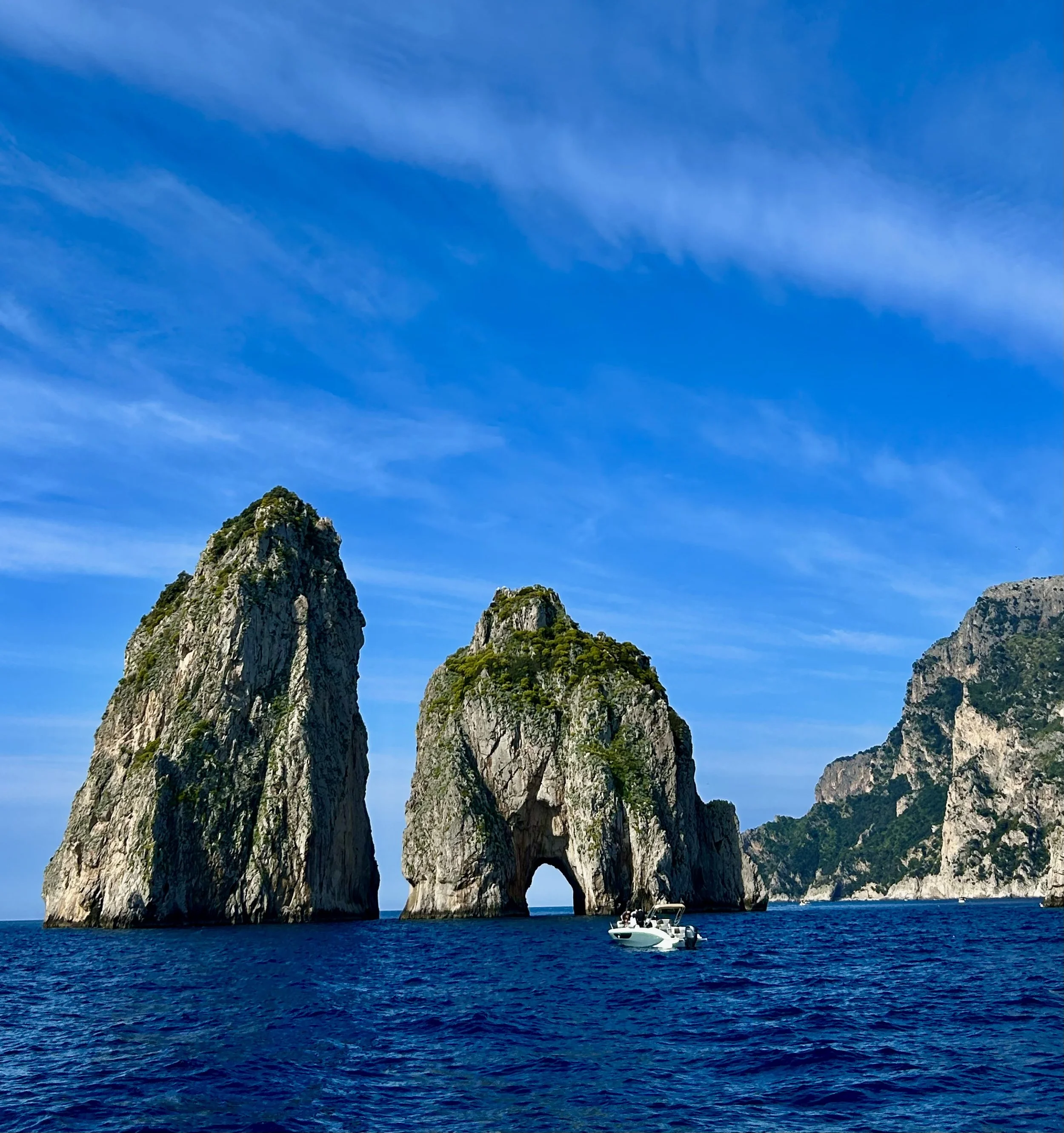 Clear blue sky with a few wispy clouds, towering limestone cliffs with arches at the shoreline, and a white boat floating on blue waters.