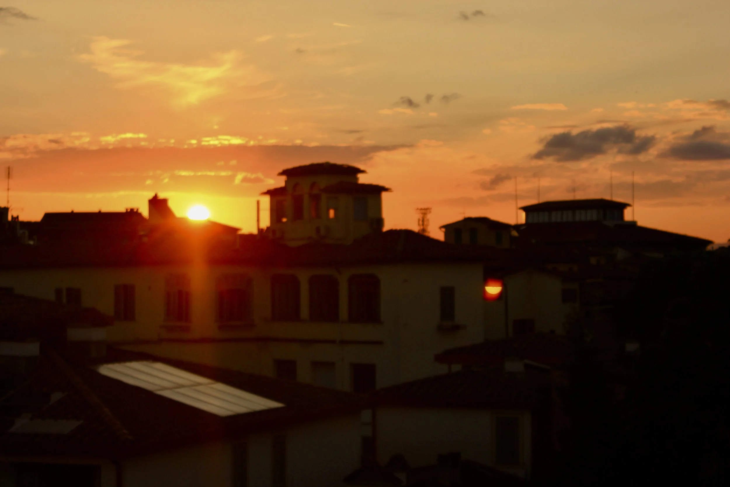 Sunset over a city skyline with buildings and rooftops, with a partly cloudy sky painted in warm hues of orange and yellow.