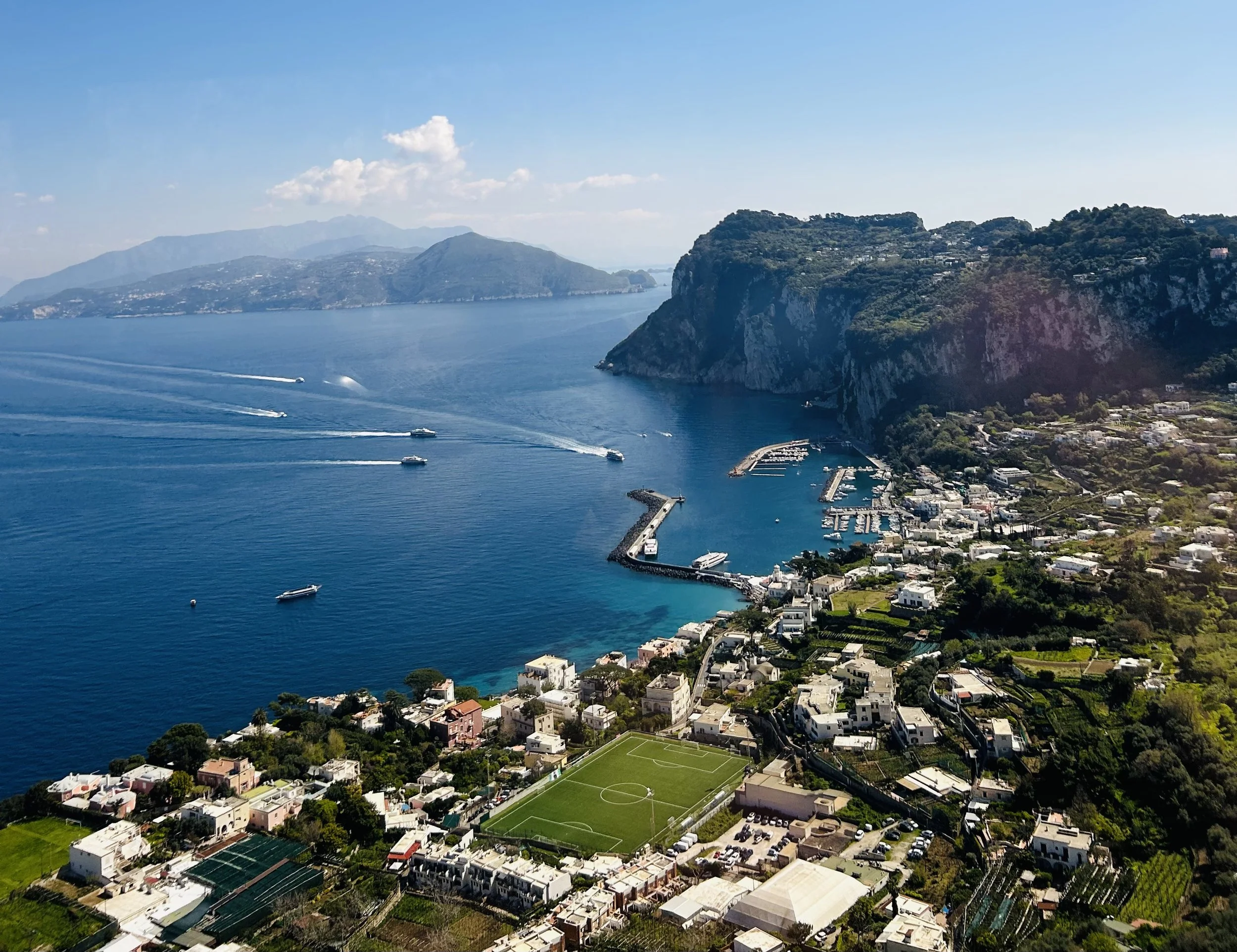 An aerial view of a coastal town with numerous white buildings, a sports field, and a marina along a large body of water. Green hills and mountains are visible in the background under a blue sky with some clouds.