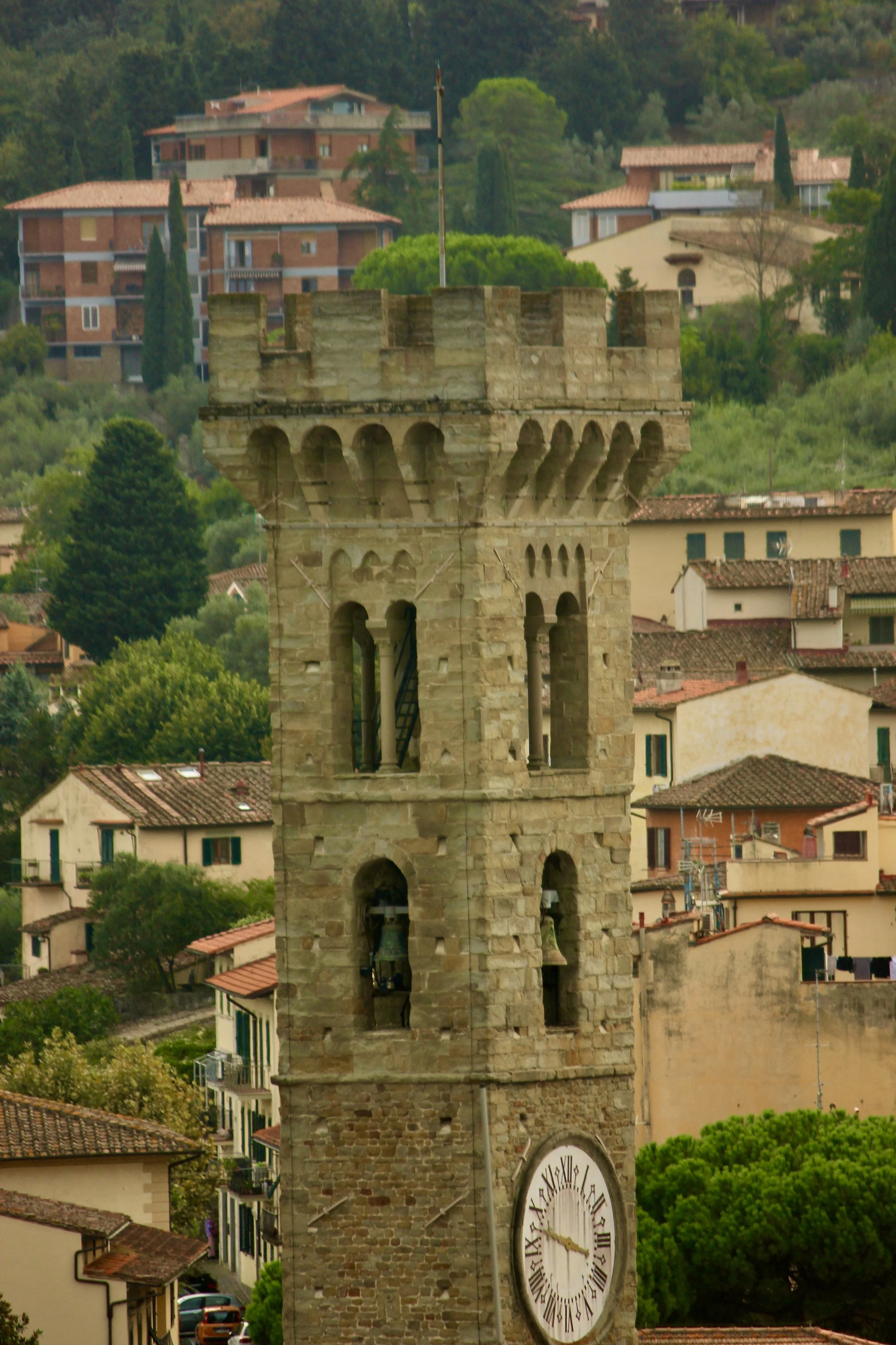 An old stone clock tower with Roman numerals, showing the time as approximately 4:07, located in a small town with houses on a hillside surrounded by green trees.