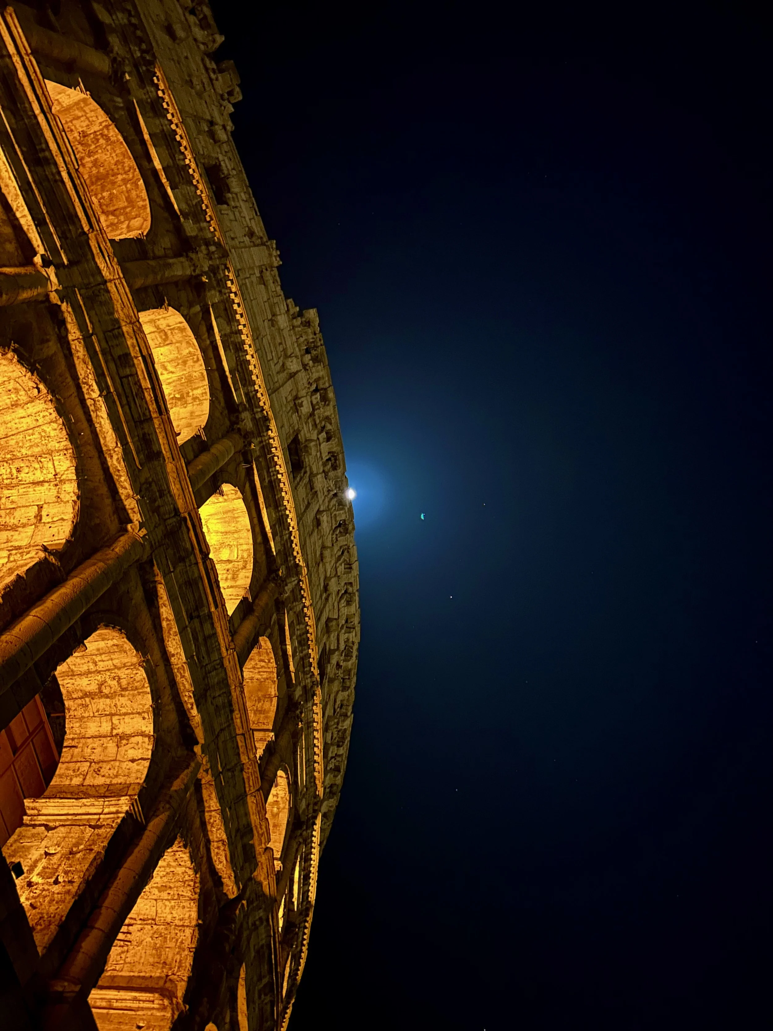 Nighttime view of the Colosseum in Rome, Italy, illuminated with warm lights, against a dark sky with a few stars visible.