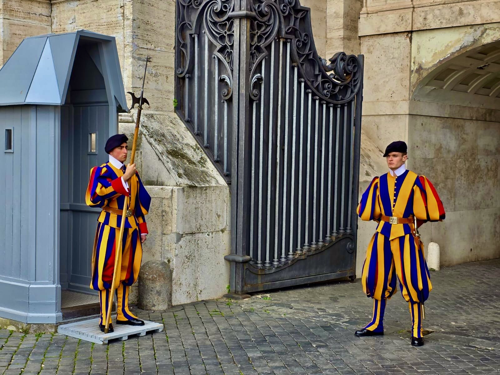 Two Swiss Guard soldiers in colorful striped uniforms standing guard near a large wrought iron gate and small guardhouse.