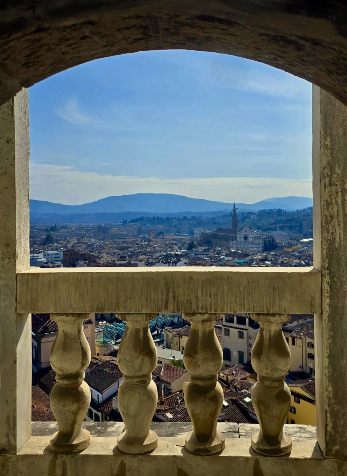 View of a cityscape through a stone window with ornate balustrades, including a church with a tall steeple, surrounded by buildings and mountains in the distance under a blue sky.