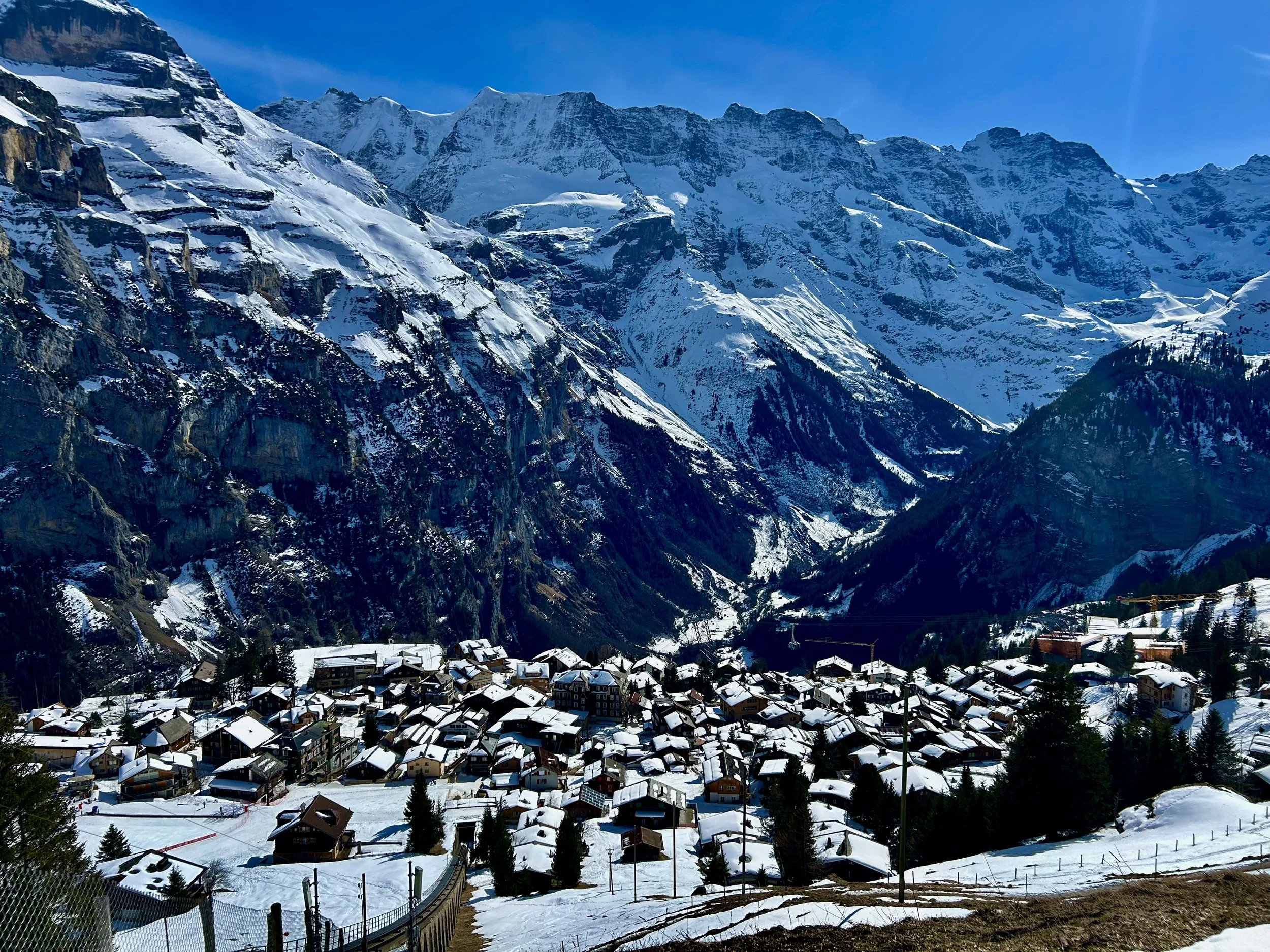 Mountain village with snow-covered rooftops and a backdrop of tall, rugged, snow-capped mountains under a clear blue sky.