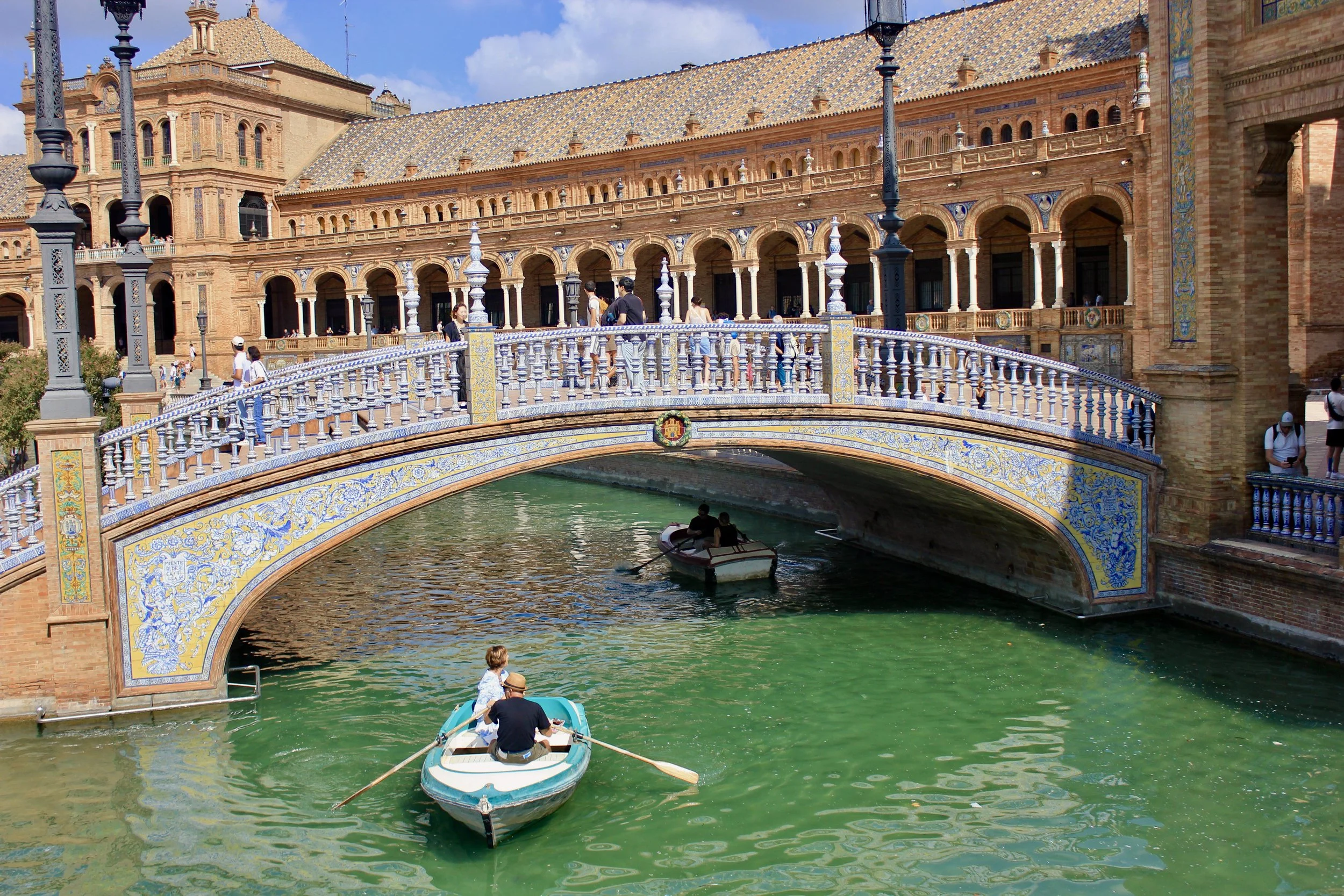 A scenic view of a historic building with a bridge and canal in Venice, Italy. The building has ornate architectural details, and there are people walking and boating in the canal.