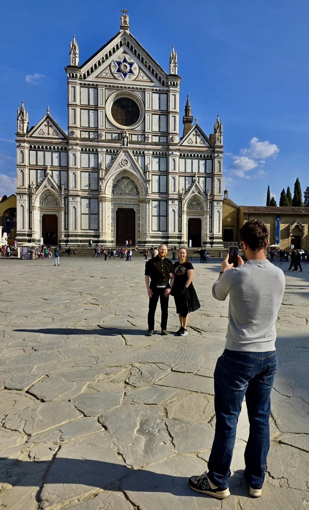 Tourists taking a photo in front of the Basilica di Santa Croce in Florence, Italy, with a clear blue sky overhead.
