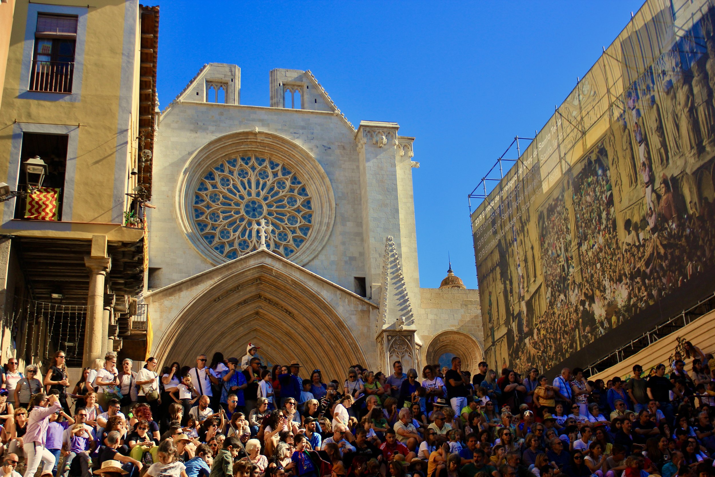 Crowd of people seated on steps outside a historic church with a large circular stained glass window, blue sky, and a modern billboard on the right.
