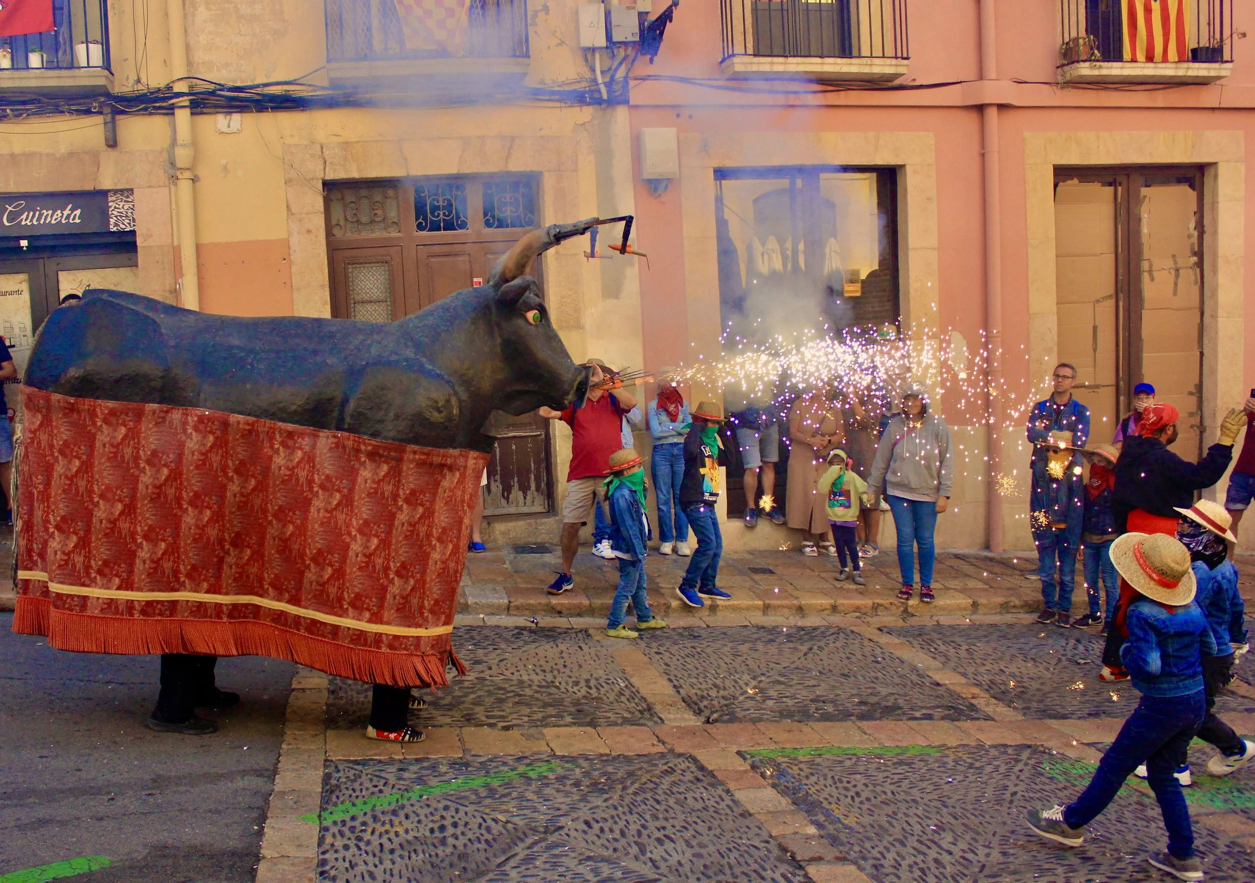 A street scene during a festival with a large bull sculpture shooting sparks, surrounded by people including children and adults, some wearing hats and taking photos, in front of colorful buildings with balconies.