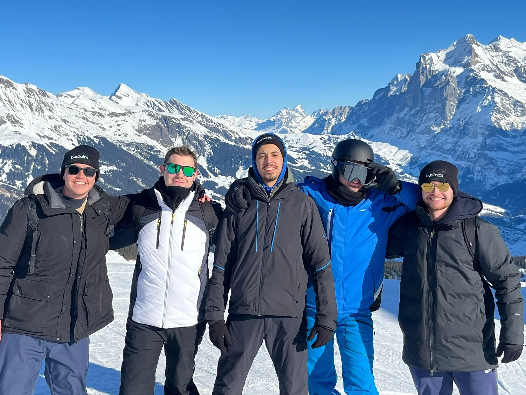 Four people dressed in winter gear standing on snow-covered ground with snowy mountain peaks in the background.