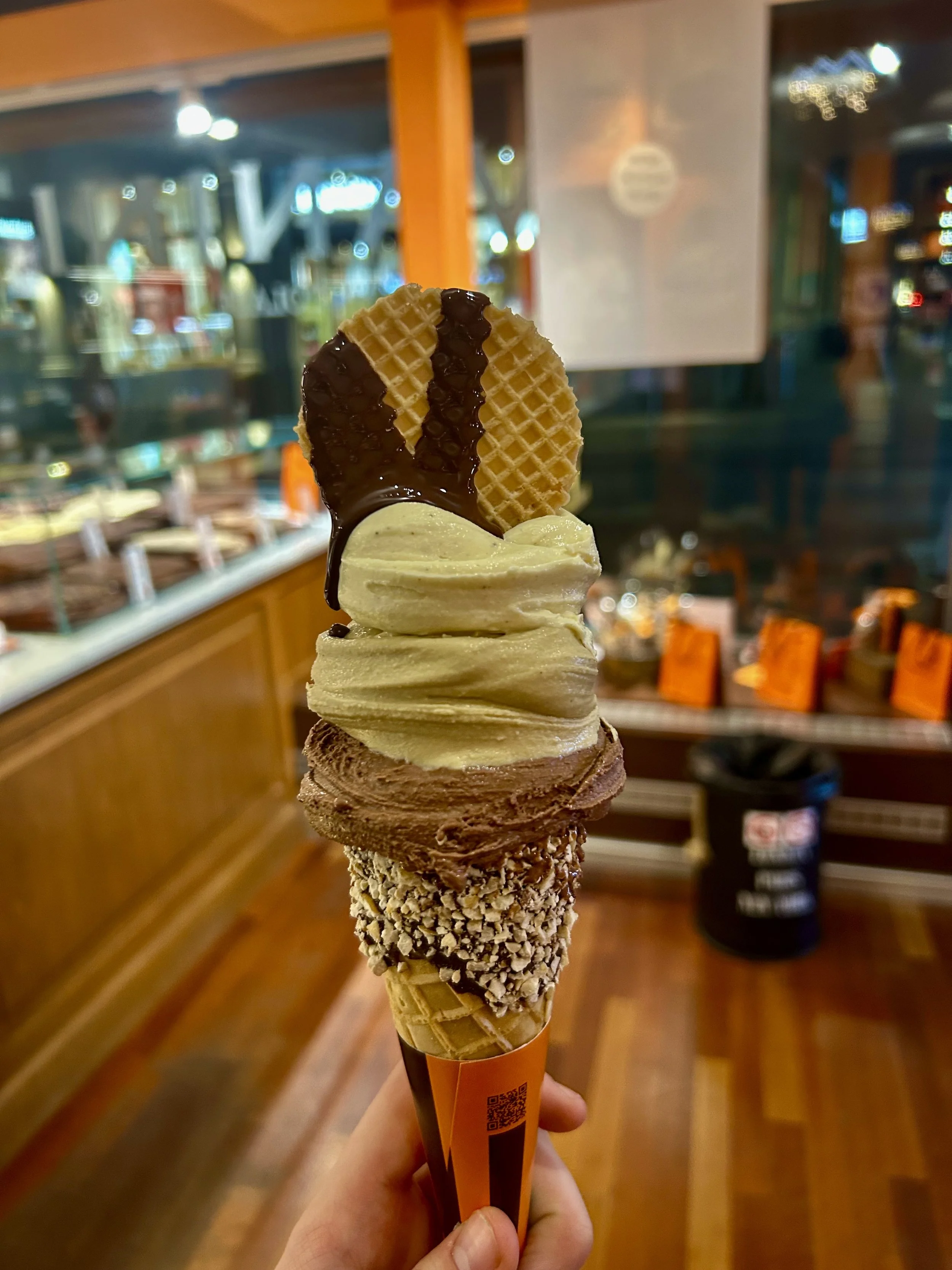 Close-up of a multi-flavor ice cream cone with various toppings, held by a person's hand inside a store with wooden furniture and bright lights in the background.