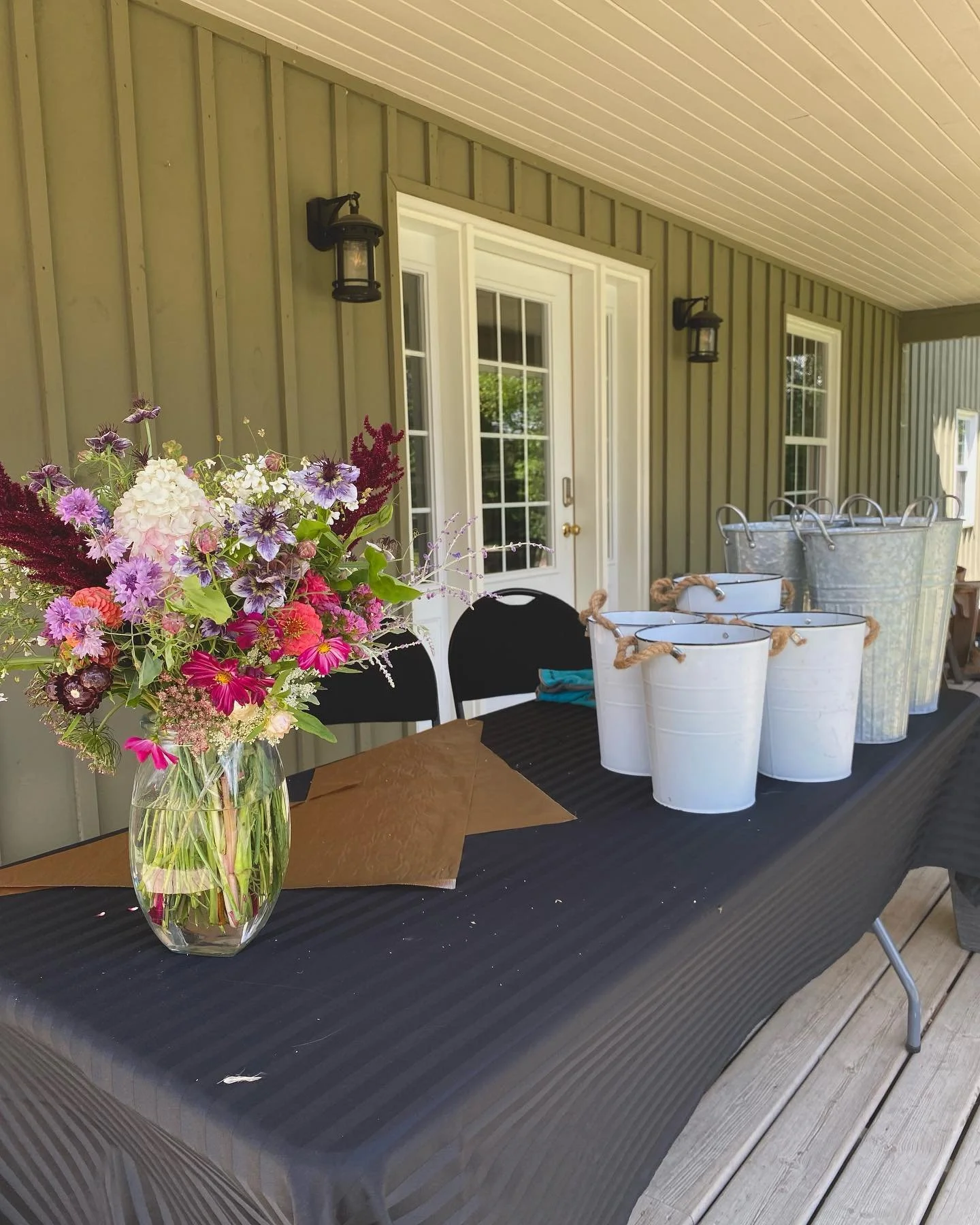 A table with a large colorful flower arrangement in a glass vase and several white buckets on a black tablecloth on a wooden deck outside a house with green siding and white door and window trim.