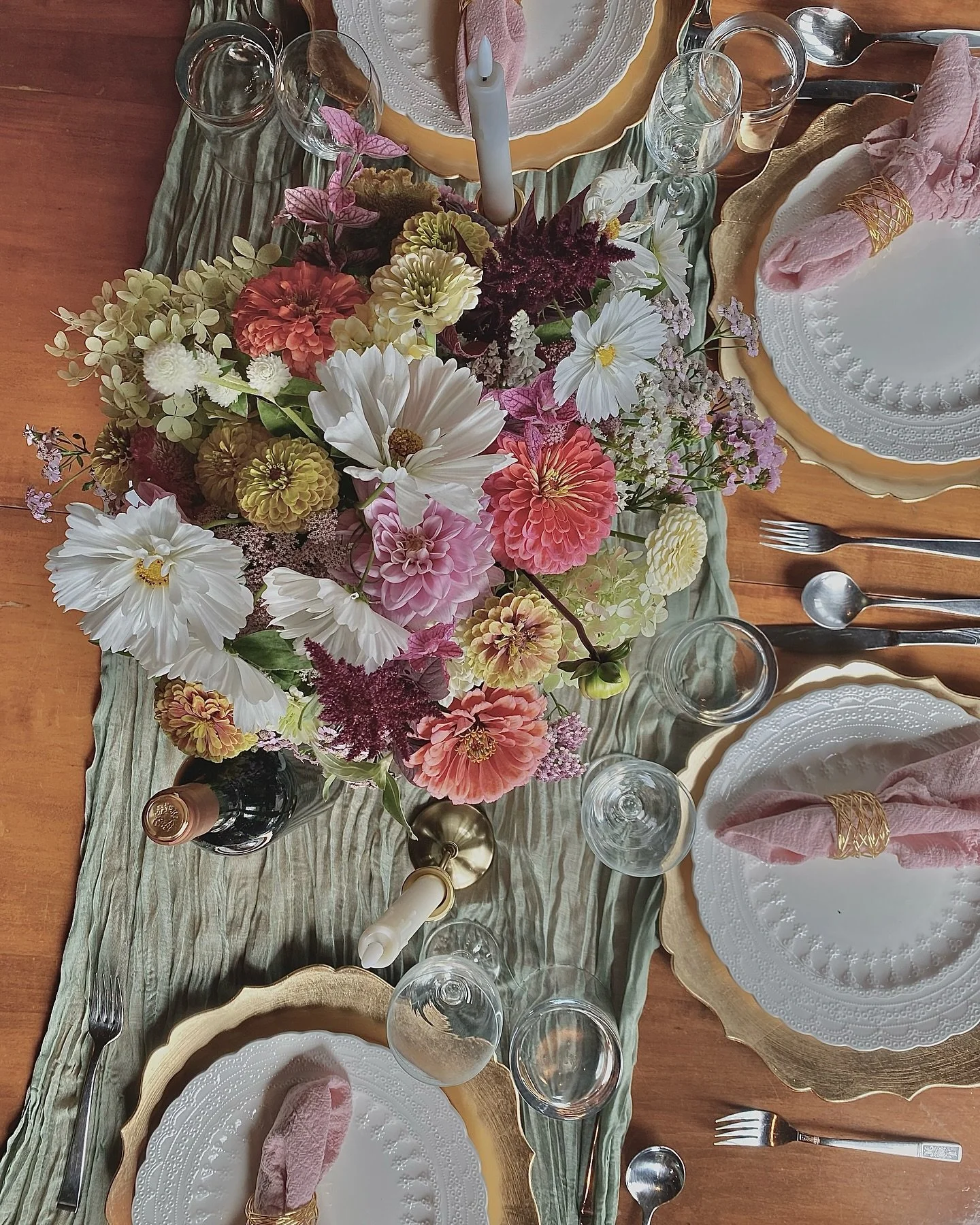A table set for a meal with a flower arrangement centerpiece, white plates with decorative gold rims, pink cloth napkins with gold napkin rings, silverware, wine glasses, and a candle. The table has a green table runner and a large yellow cake with a candle on top.