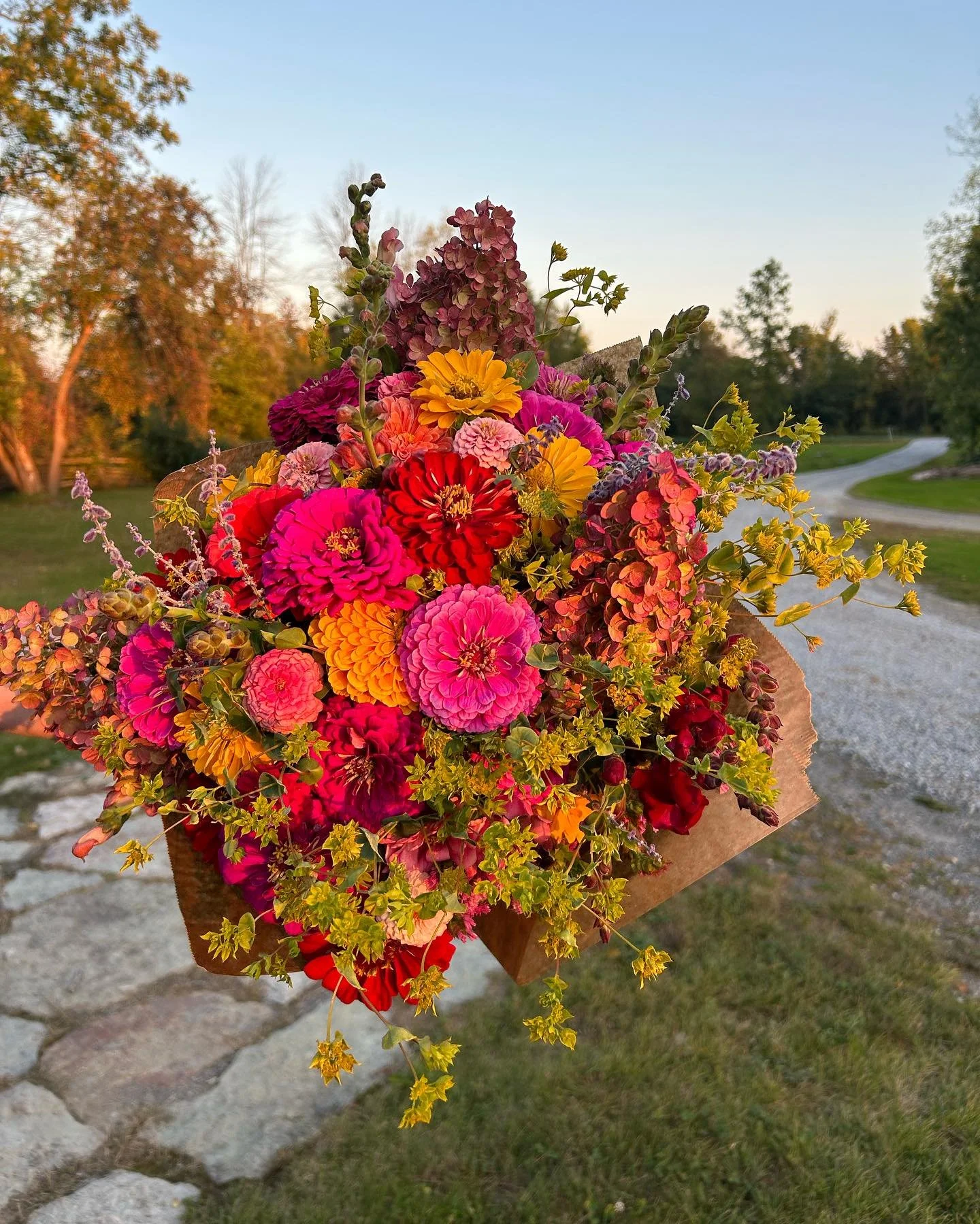 A vibrant bouquet of pink, red, yellow, and purple flowers held up with a background of trees, a dirt path, and a sunset sky.