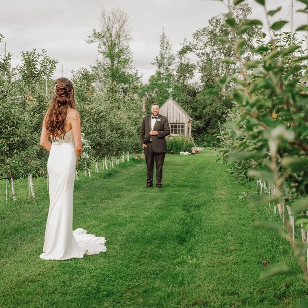 A bride and groom at their wedding outside in a lush green orchard. The bride has long hair and is wearing a white wedding gown, and the groom is in a black suit with a bow tie, smiling at her.