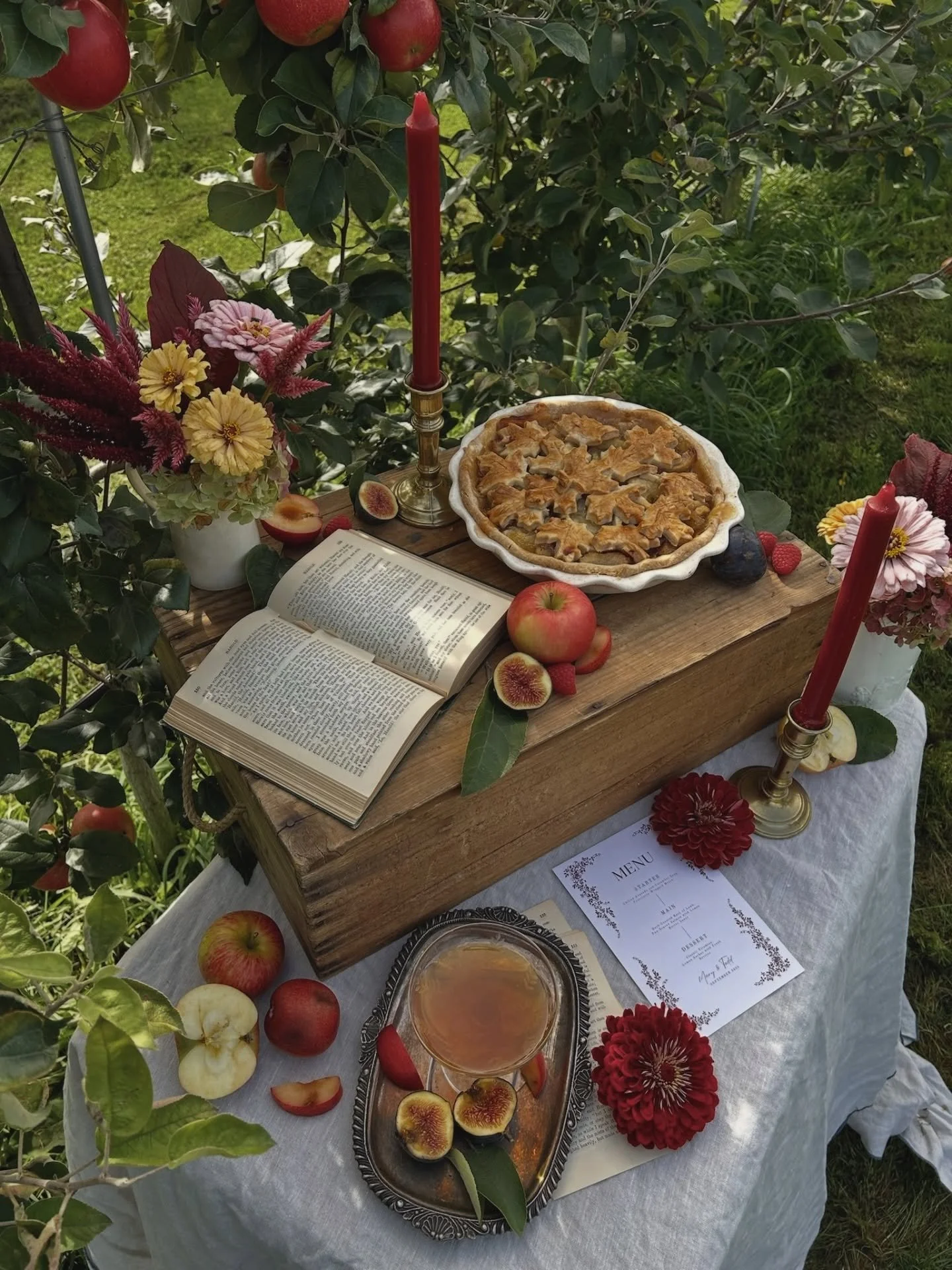A rustic wooden table set outdoors with a freshly baked apple pie, whole and sliced apples, a glass of apple juice, a menu, an open book, and floral arrangements with red, pink, and yellow flowers. Two red candles are on either side of the pie, and there is greenery in the background.