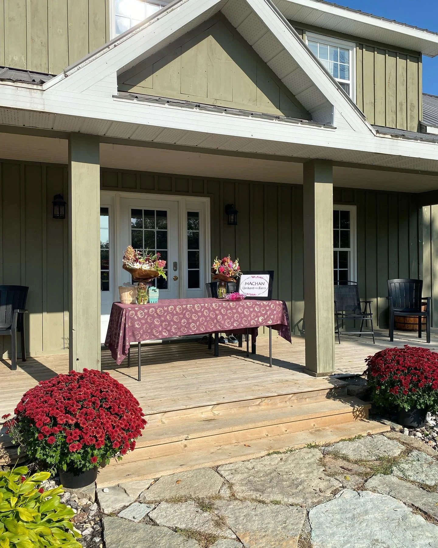 A house exterior with a wooden deck, decorated with red potted flowers and a table covered with a purple tablecloth, holding flower arrangements and a sign that reads 'MACHAN Orchard and Farm'.