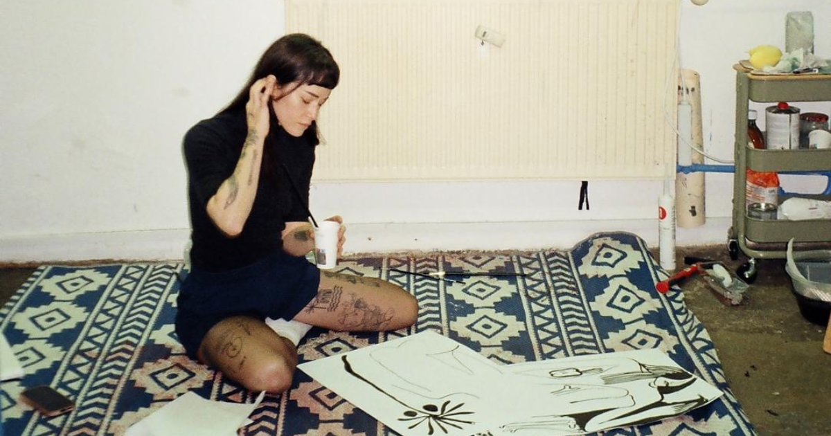 A woman with dark hair and tattoos sitting on a patterned rug in a room, holding a cup and a paintbrush, with a large drawing or painting in front of her.