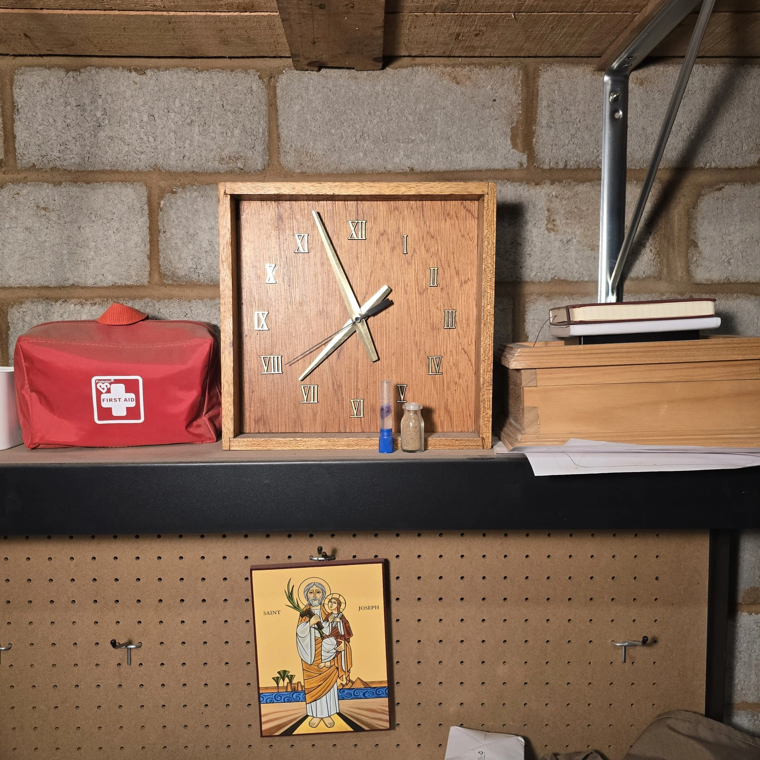 A wooden clock with Roman numerals hangs on a brick wall above a small shelf with books, a red first aid kit, and religious and decorative items.